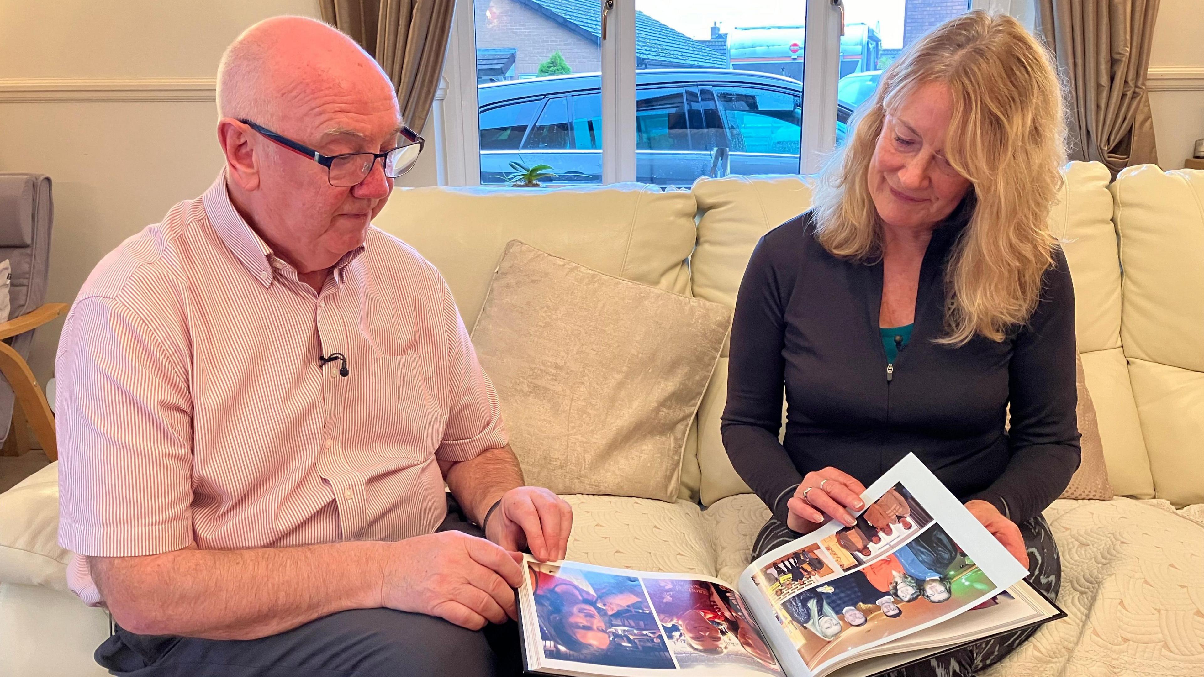 Peter McGenity is wearing a striped shirt and glasses and is sat next to Helen Price, Liam's mum, who is wearing a black cardigan. The pair are sat on a cream sofa looking through a photo book together.