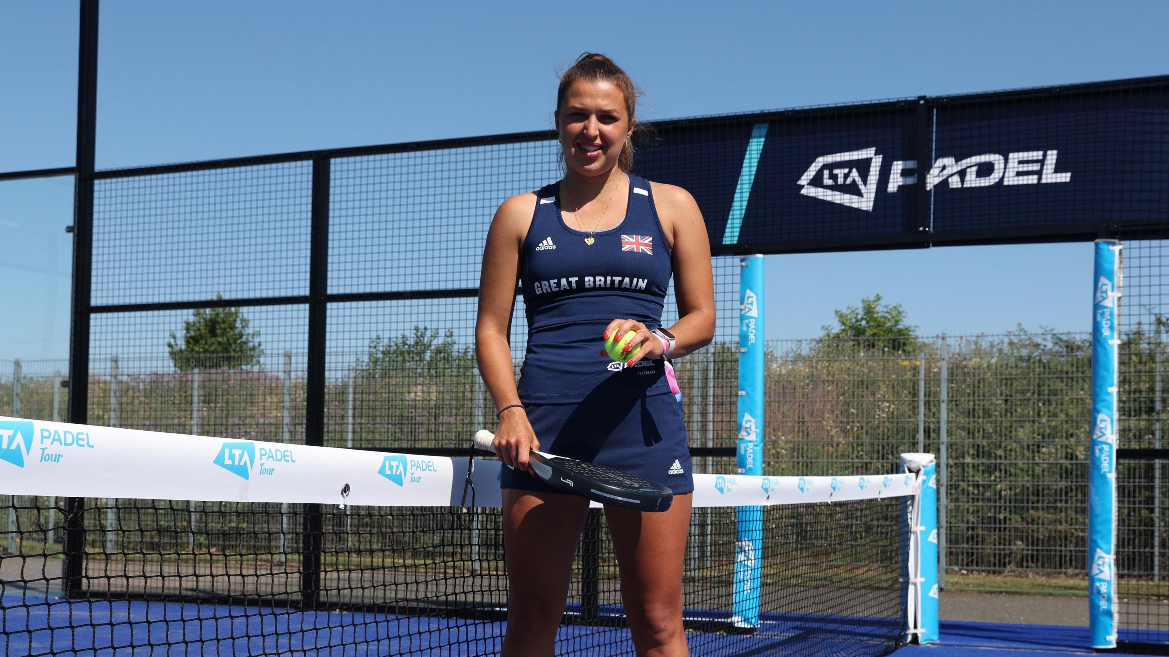 A young woman dressed in a Great Britain team vest and skirt stands next to the net in the centre of an outdoor padel court on a cloudless, sunny day. She holds her racquet at waist level and a yellow ball in her other hand.