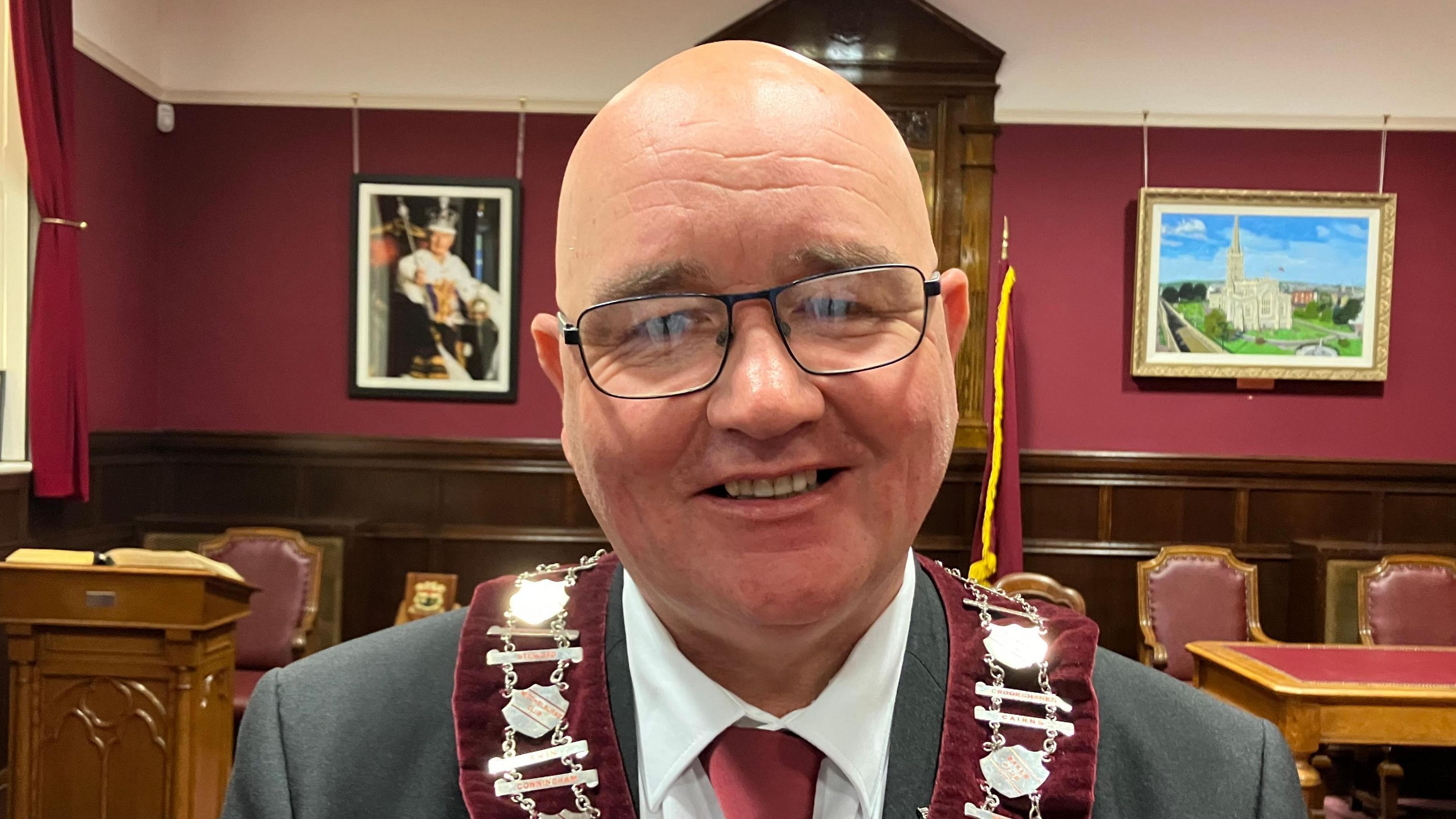William Walker is smiling and wearing glasses.
He is wearing a red collarette and a grey suit with a red tie.
