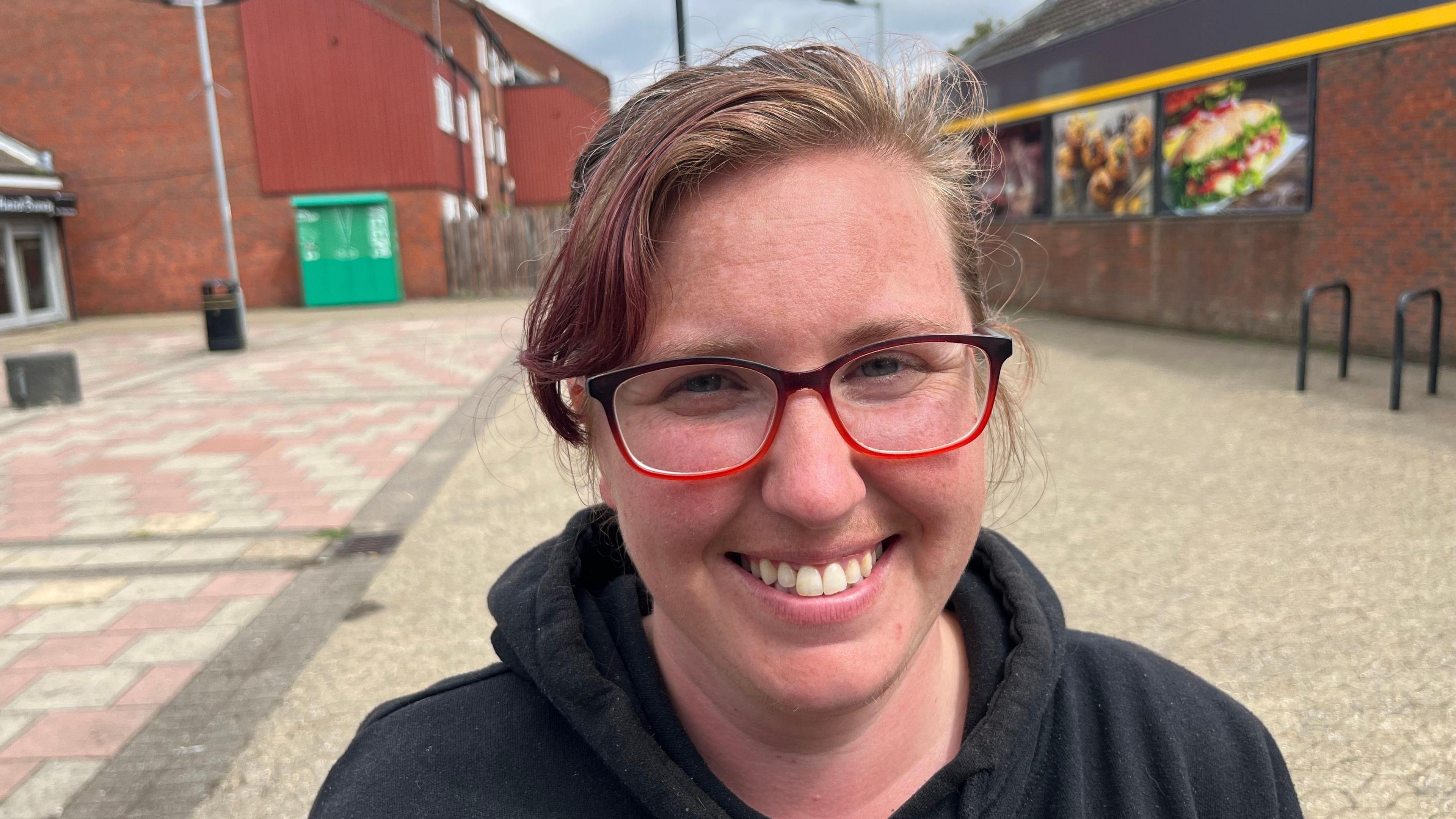 Emilia Jeeves is looking at the camera and smiling. She is wearing red and black framed glasses and a black hooded jumper. Behind her is pavement and a fast food restaurant.