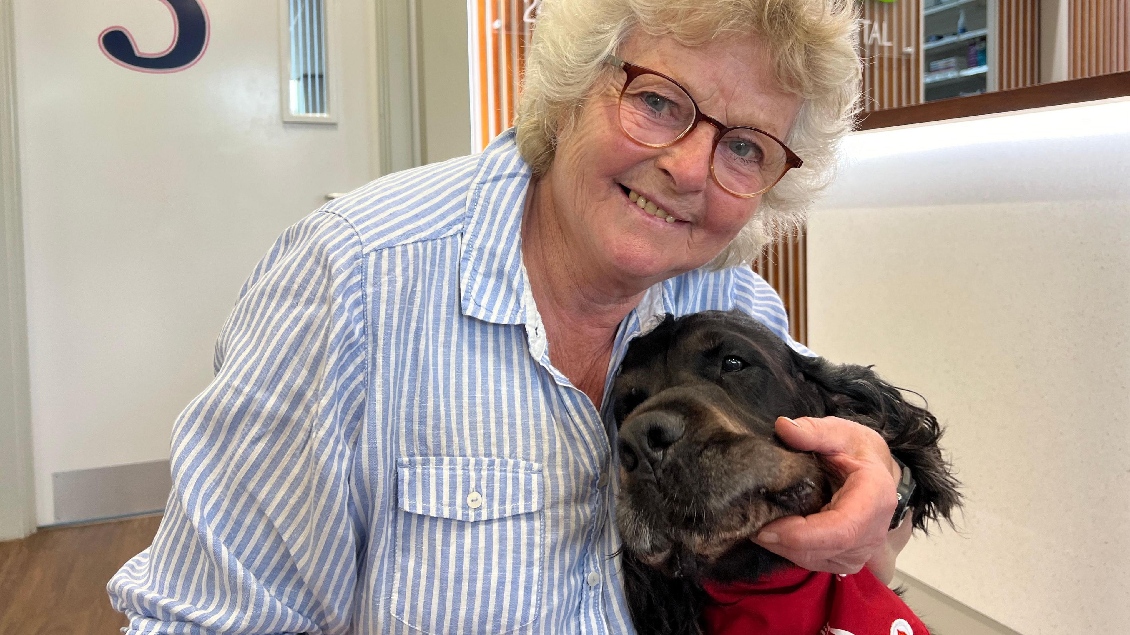 A woman with curling light coloured hair and wearing glasses cuddling her dog's head 