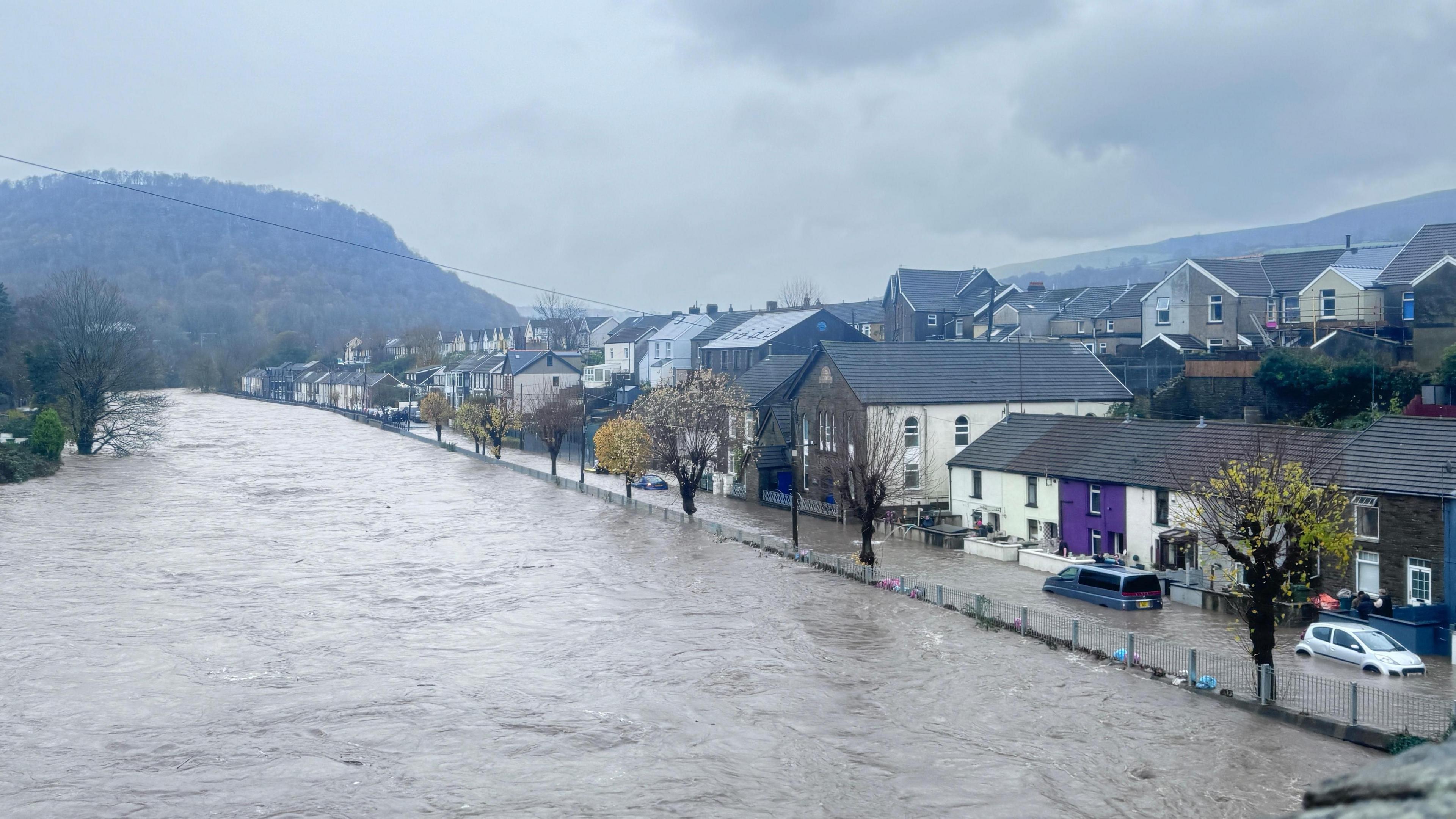 Wide image of River Taff in Pontypridd, showing the river having burst its banks. Floodwater can be seen in a street directly to its right, submerging cars in water.