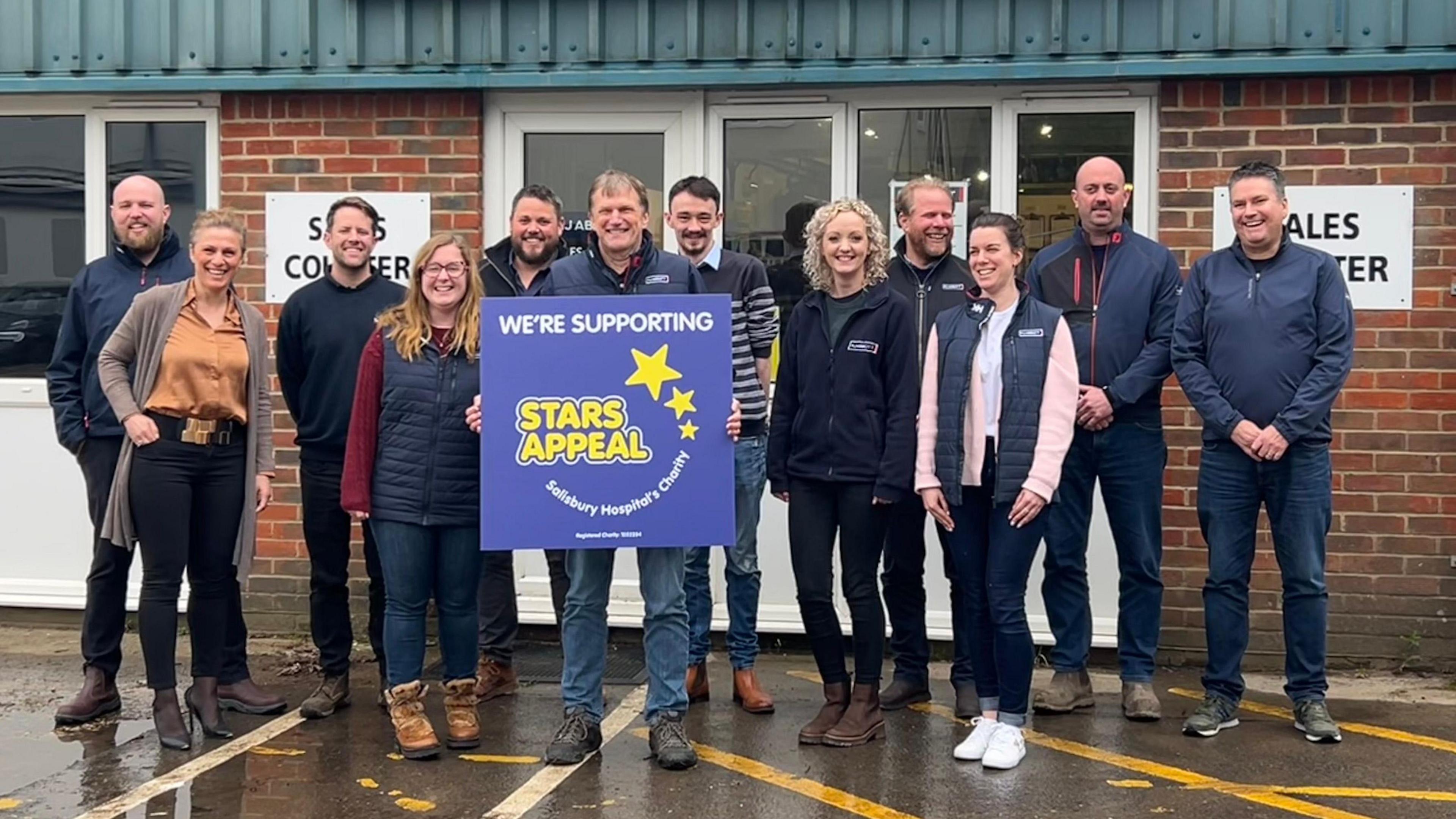 Adrian Abbot holding up a blue sign that says 'We're supporting Stars Appeal'. He is standing in a line alongside eleven other colleagues from MJ Abbott against a brick wall. 