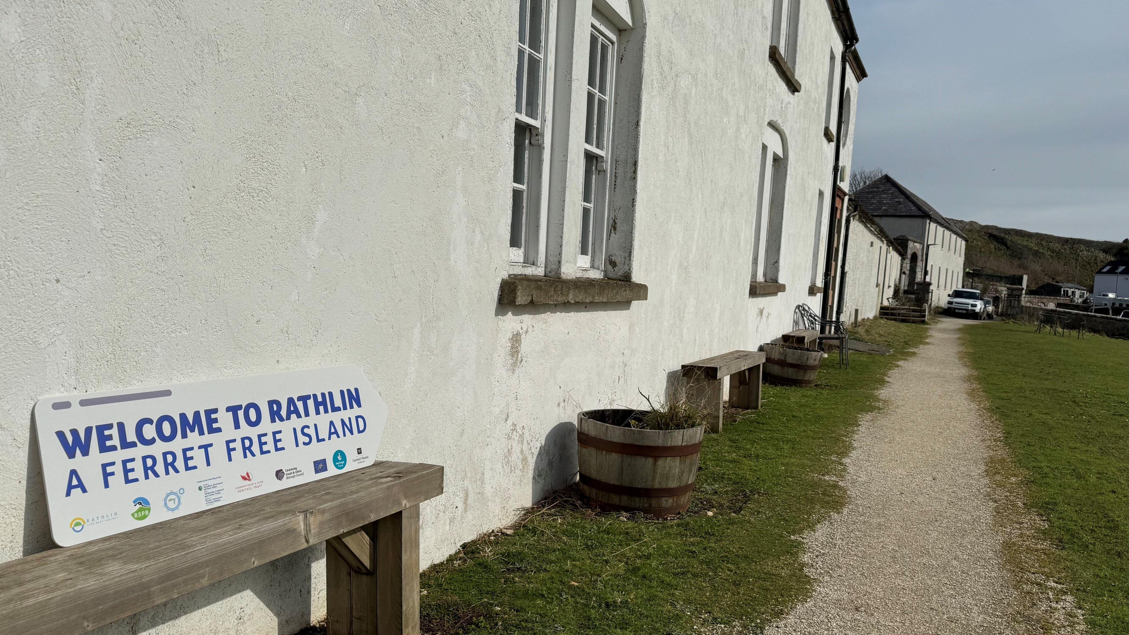 A sign says welcome to Rathlin, a ferret free island. It's been placed on a wooden bench next to a house. 