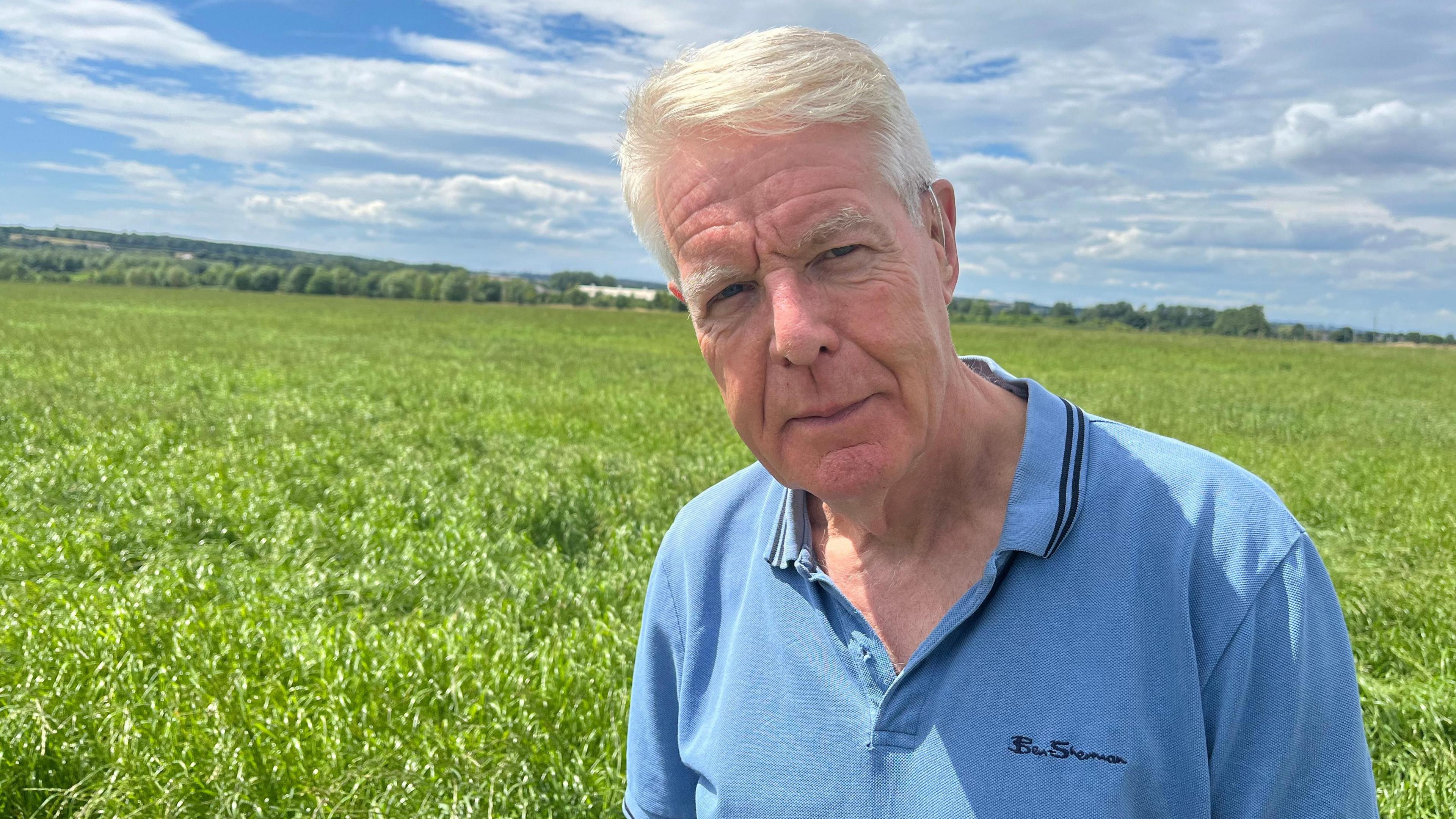 A white-haired man in a blue polo shirt standing in front of a green field