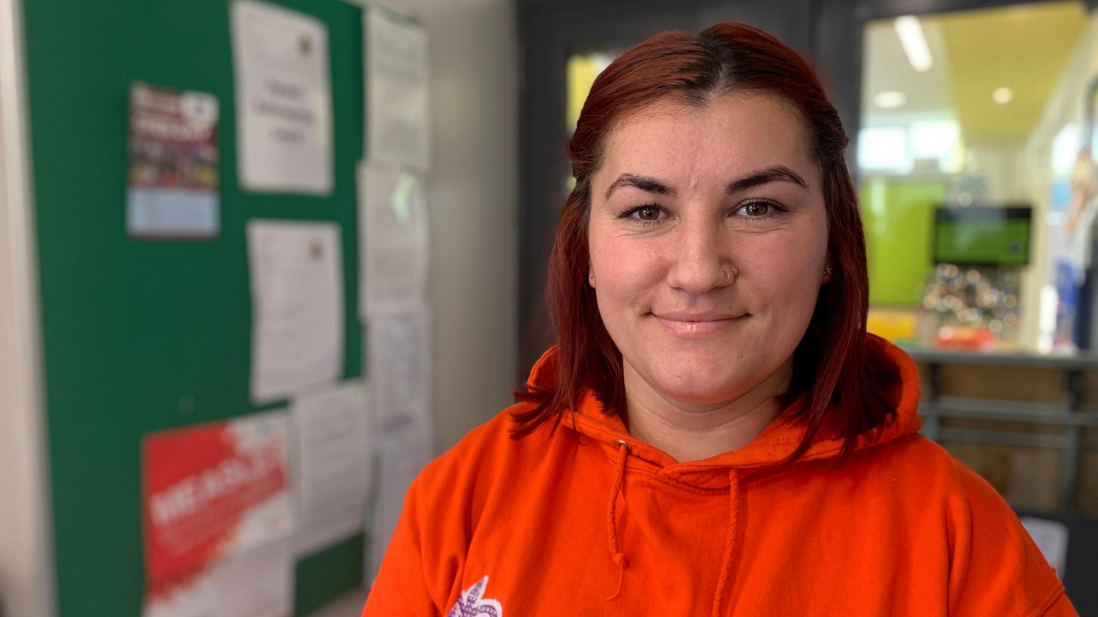 Ms Pintea with shoulder length brown hair, she is wearing an orange hoodie as she smiles at the camera. 