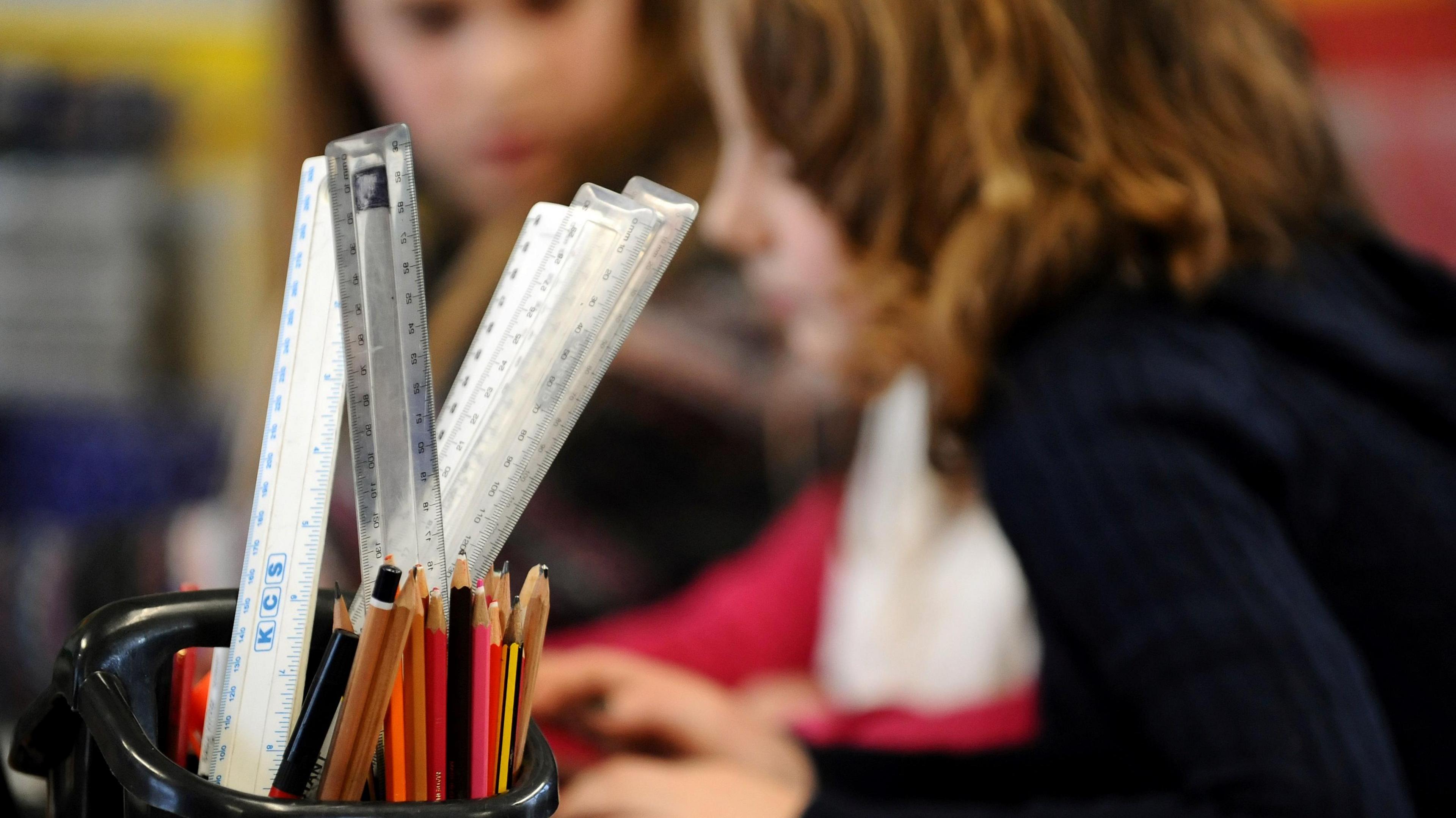 A stock image of a pen holder with pencils and rulers in the foreground, with a blurred background showing two girls sat doing homework