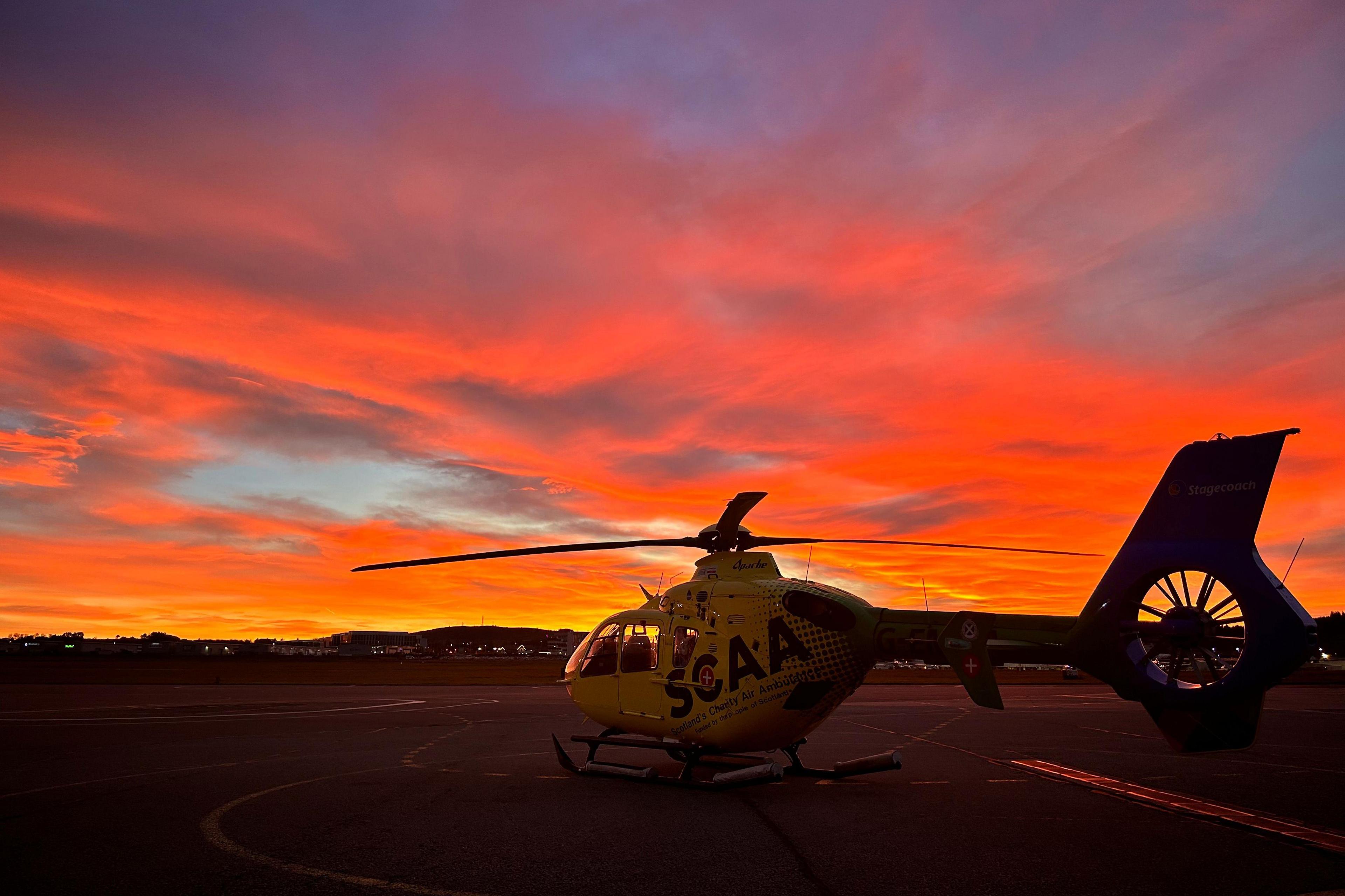 A yellow helicopter sits on the tarmac with a sunset behind it. The sky is orange, red and yellow.