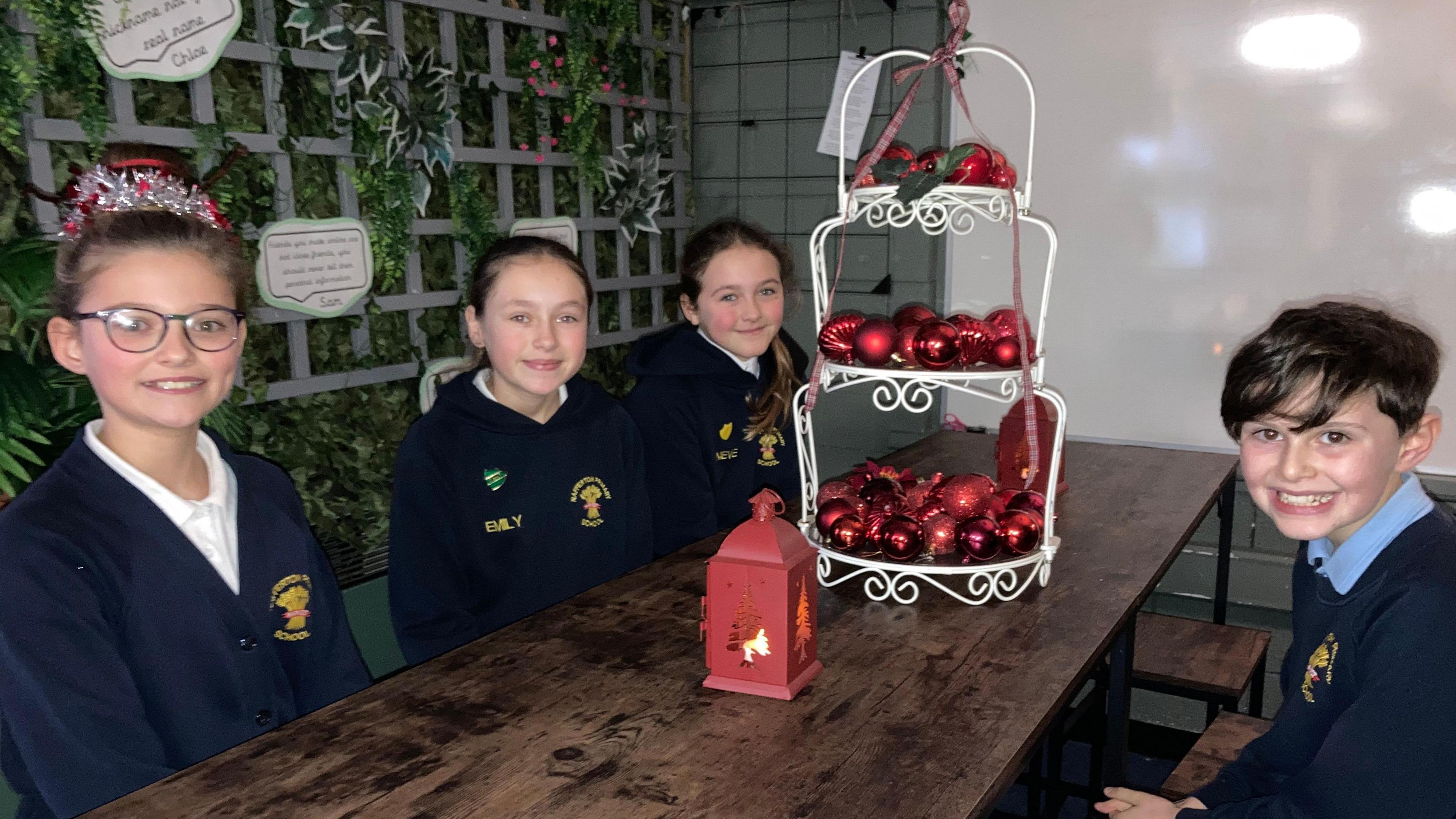 Left to right, Alice, Emily, Nieve and Jonah sat on a wooden bench. On the wooden table is red Christmas baubles and a small red Christmas decoration with a light inside. All students are sat smiling looking into the camera.