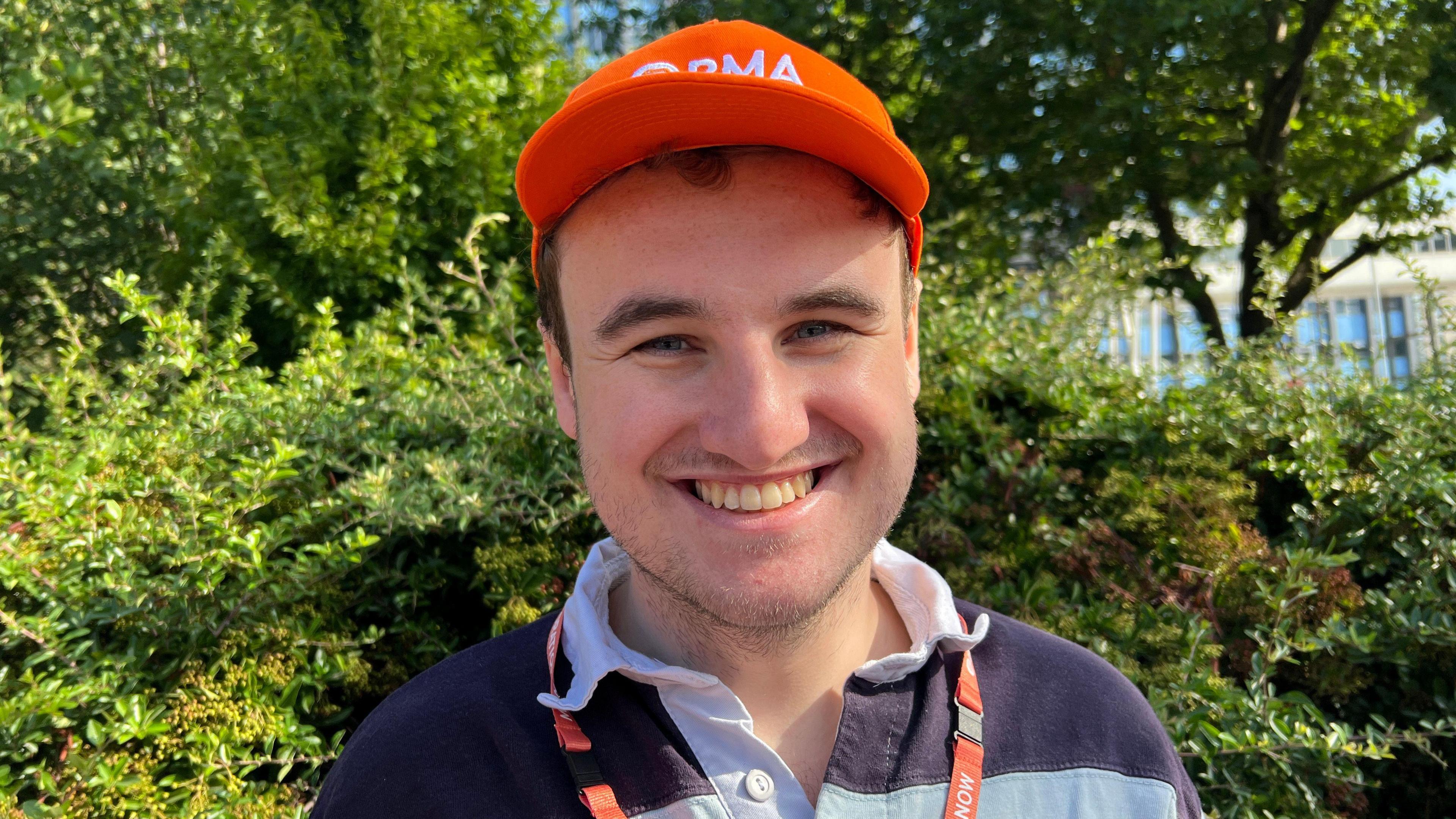 A man with short hair is wearing an orange BMA cap and an orange lanyard with a navy and light blue polo shirt. Behind him are trees and there is a building in the distance.