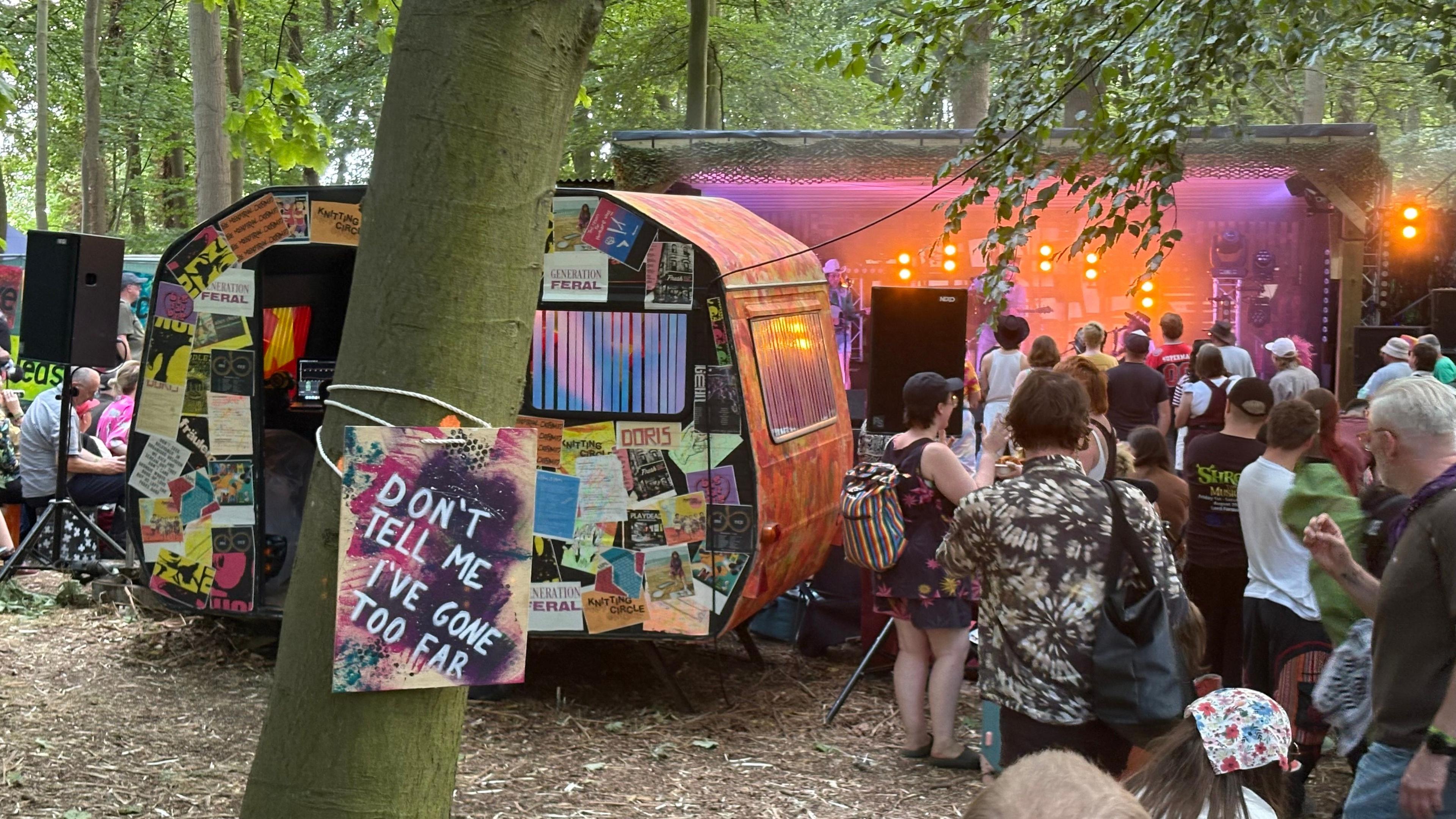 A colourful caravan covered in posters is parked behind a tree on which is tied a notice saying "don't tell me I've gone too far".  There is a stage bathed in pink light in the background and a large crowd of people.