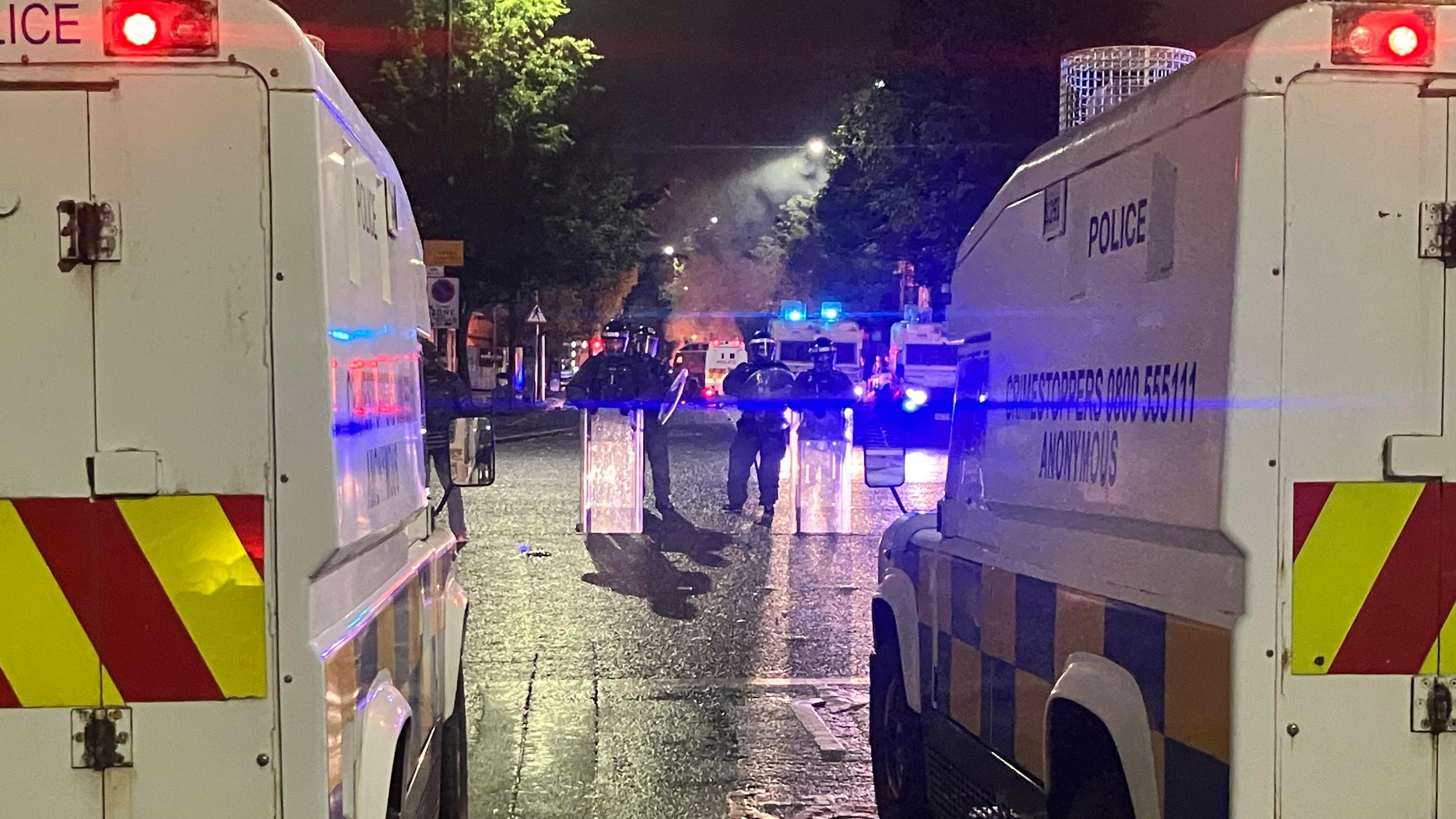 Police wearing riot gear and holding shields with two riot vans are parked on either side