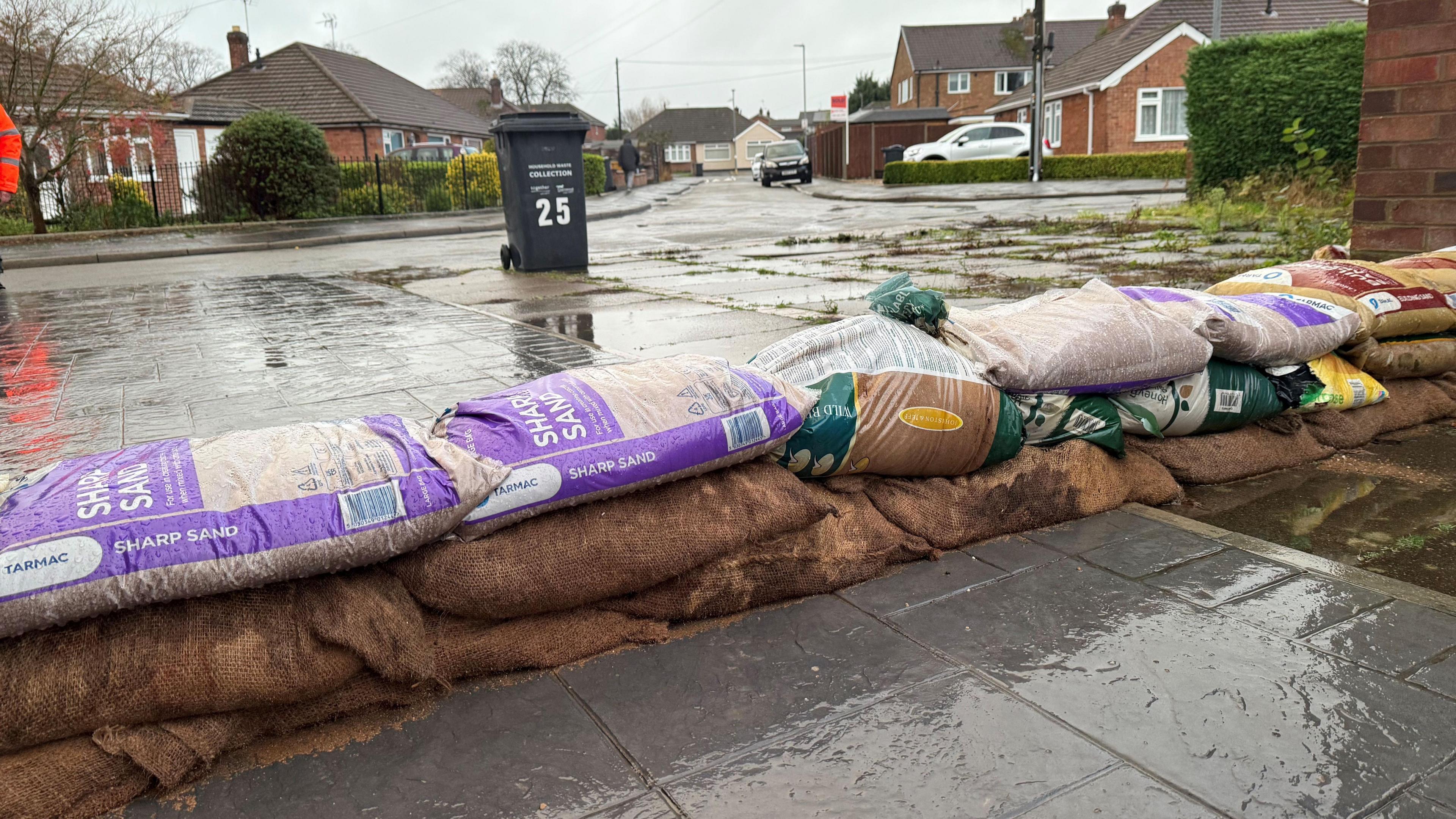 A row of sandbags stacked across a paved area, with a residential street in the background.