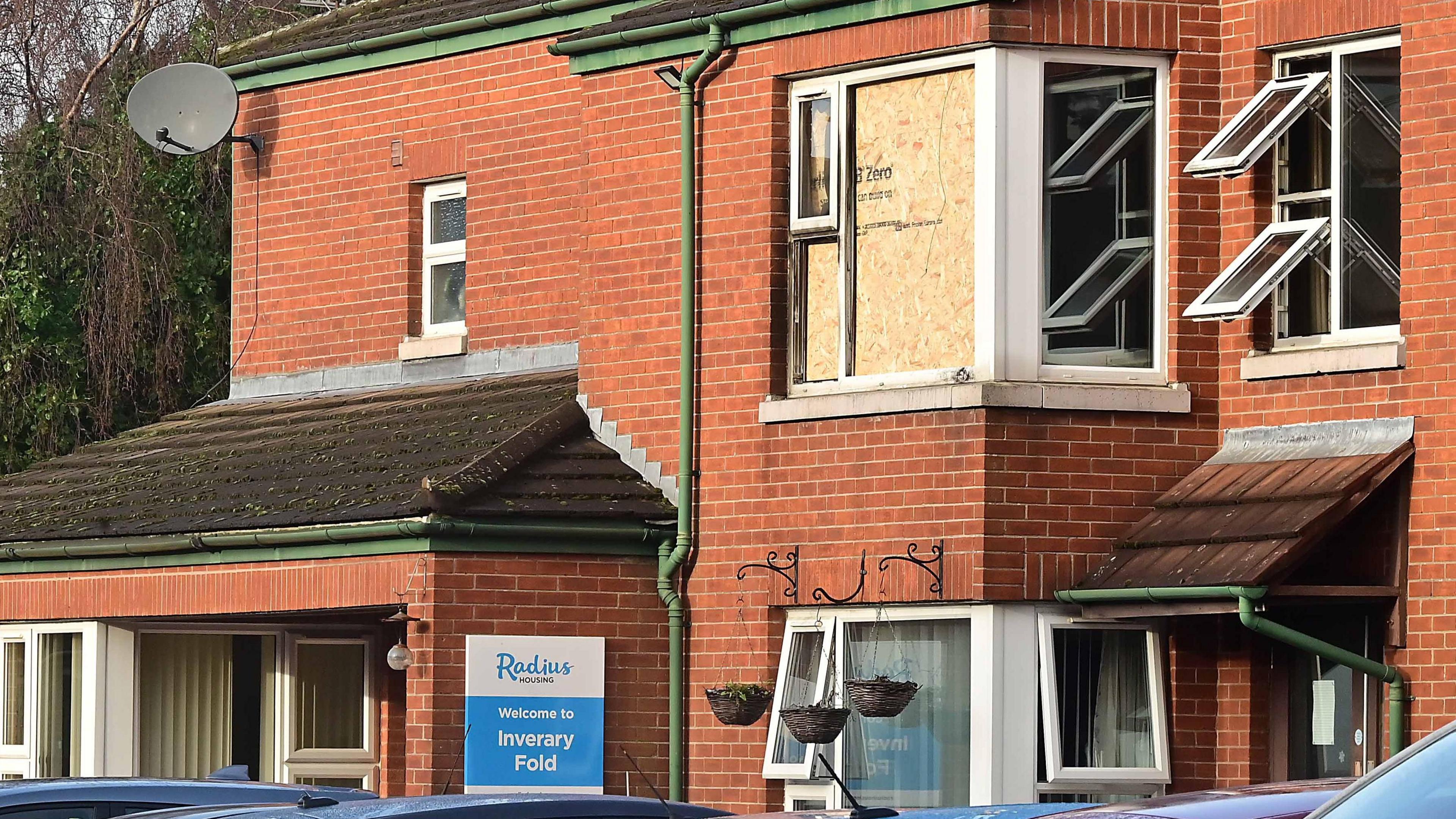 A corner window on the first floor of a red-brick building is boarded up with plywood. There is a blue and white sign on the ground floor exterior wall that says 'Radius Housing: Welcome to Inverary Fold'.