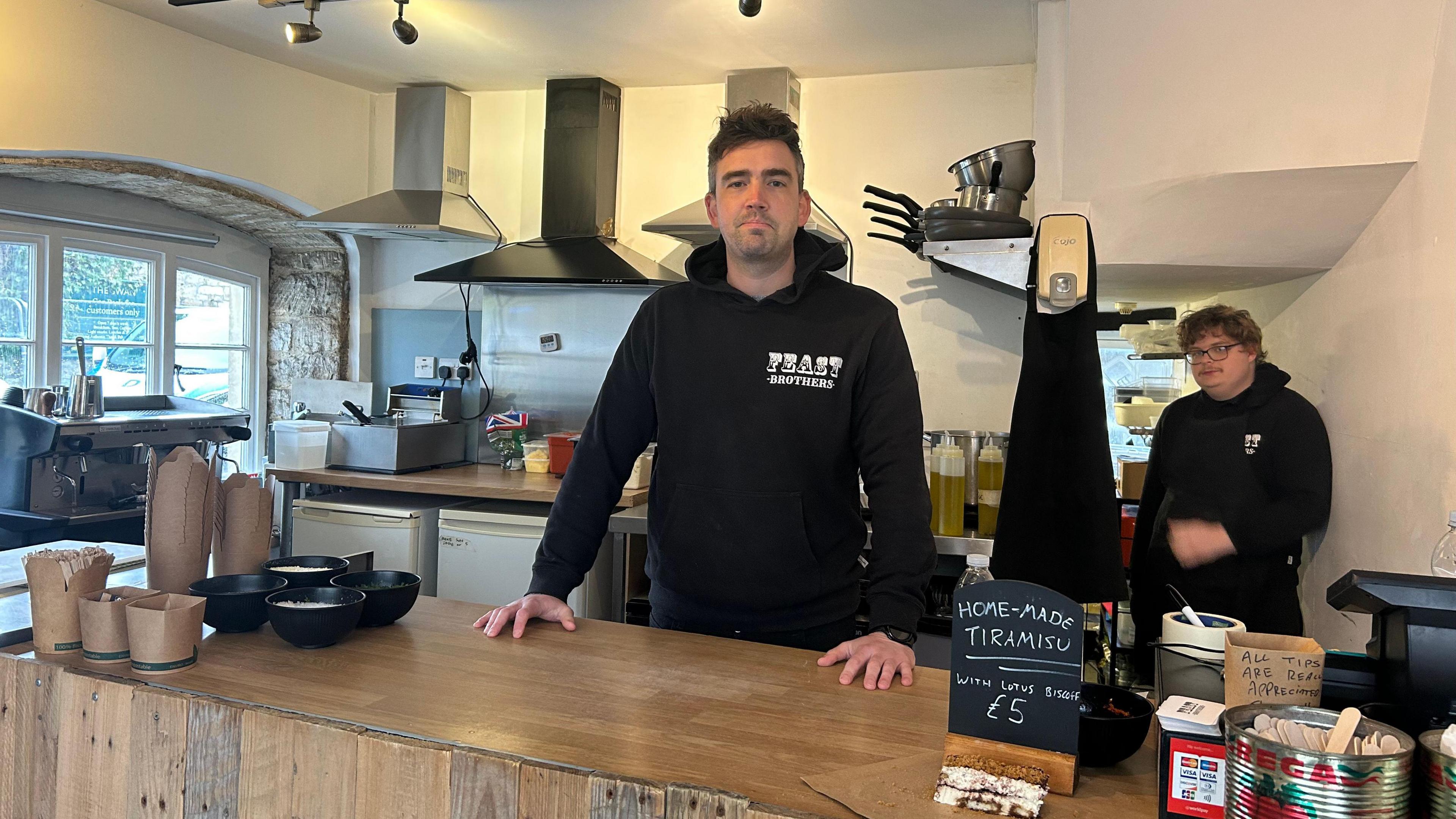 Scott Wheeler standing behind the counter of his business called Feast Brothers in Bradford-on-Avon. He is wearing a black hoodie and has dark hair