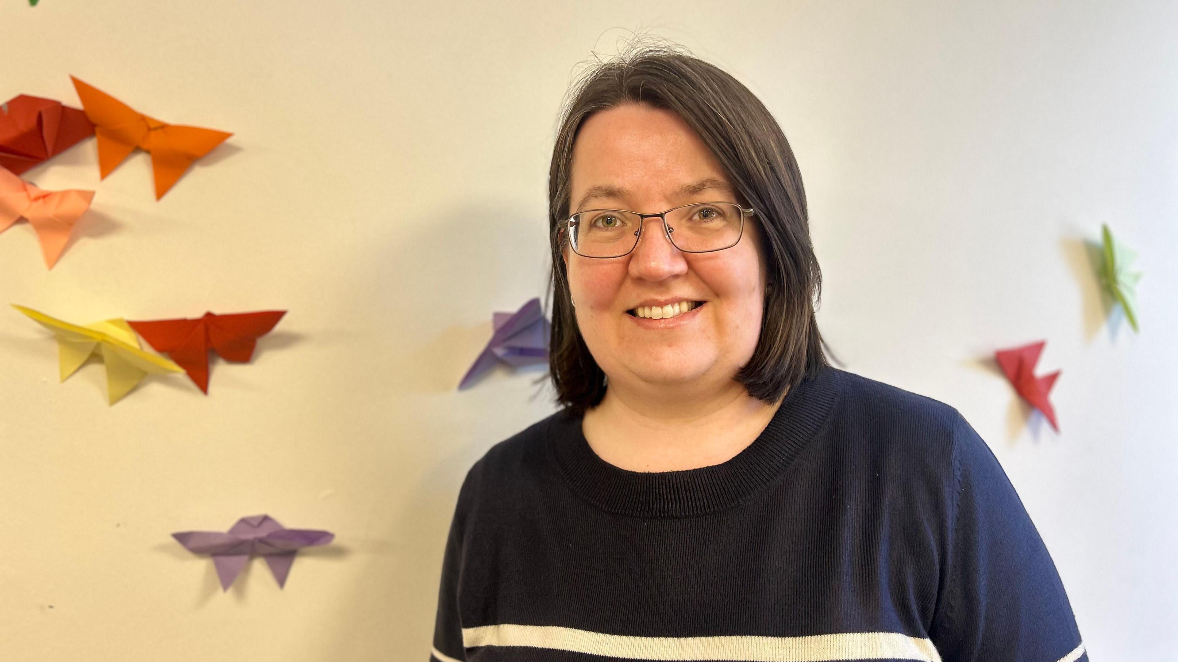 A lady with glasses and short hair smiling next to a wall of origami butterflies