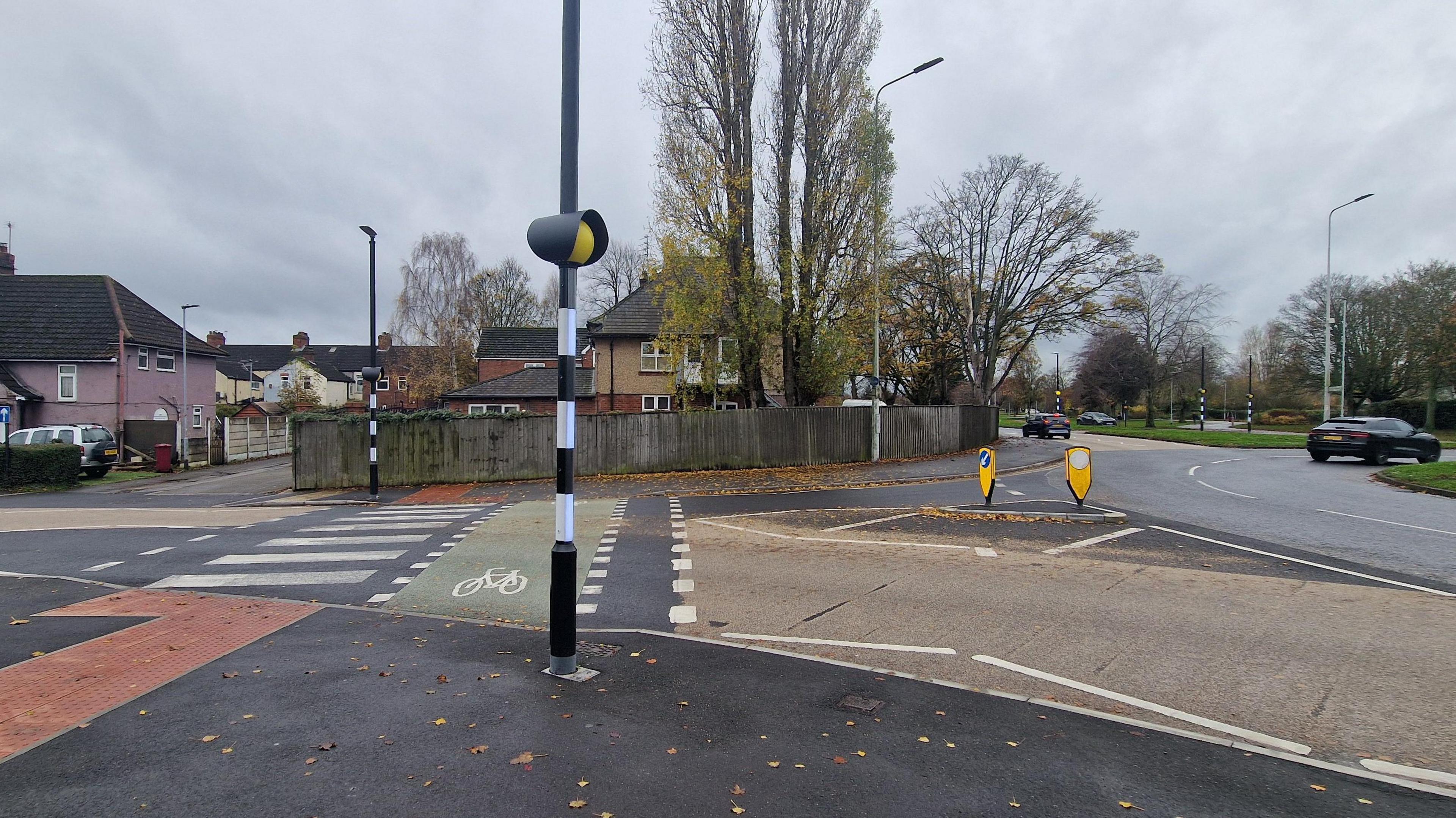 Zebra crossing near a roundabout with another zebra crossing in the distance. There are residential buildings / houses and two cars as well as trees in the background.
