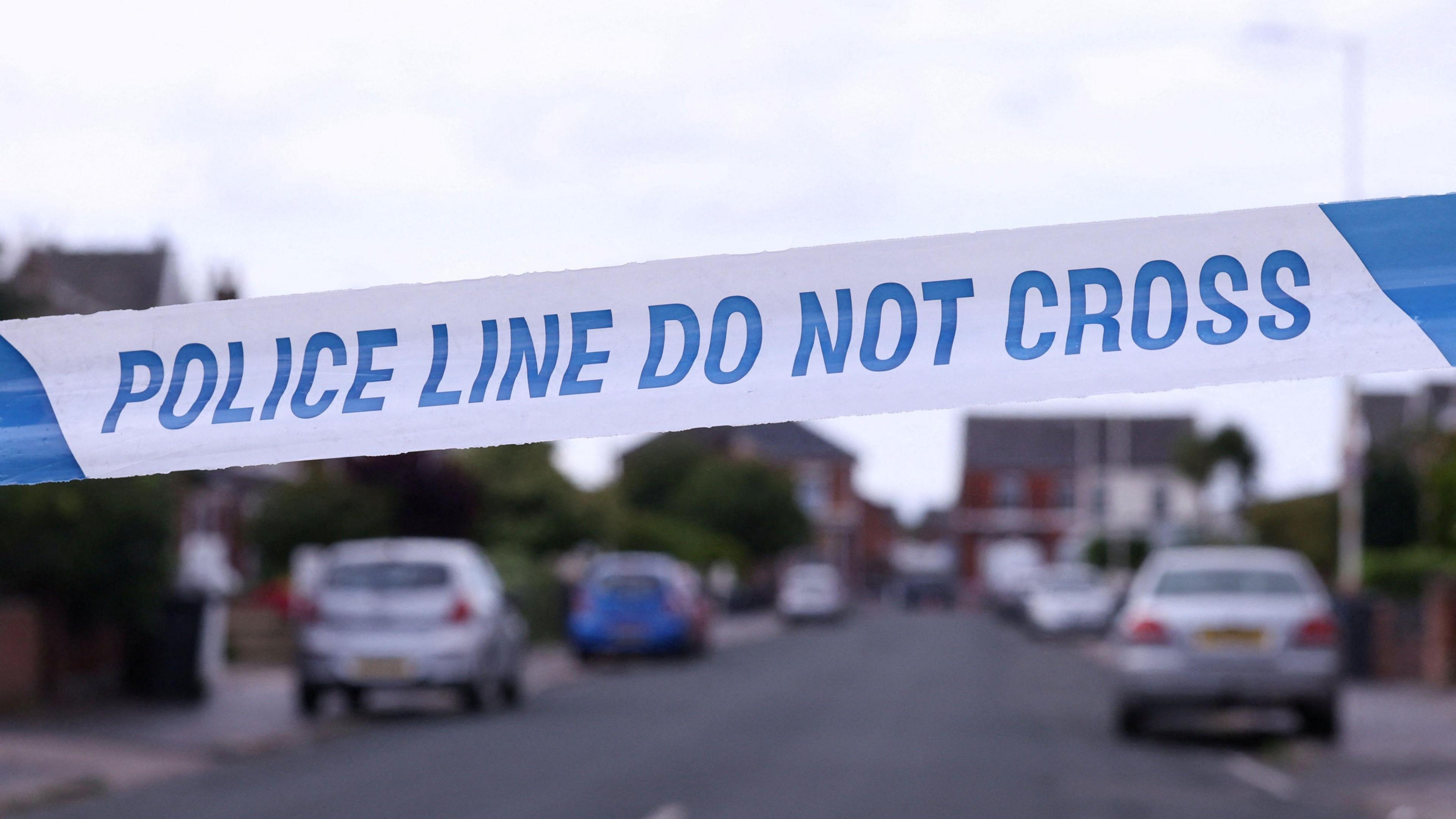 A close-up shot of blue and white police tape with the words "police line do not cross" with a residential street in the background.