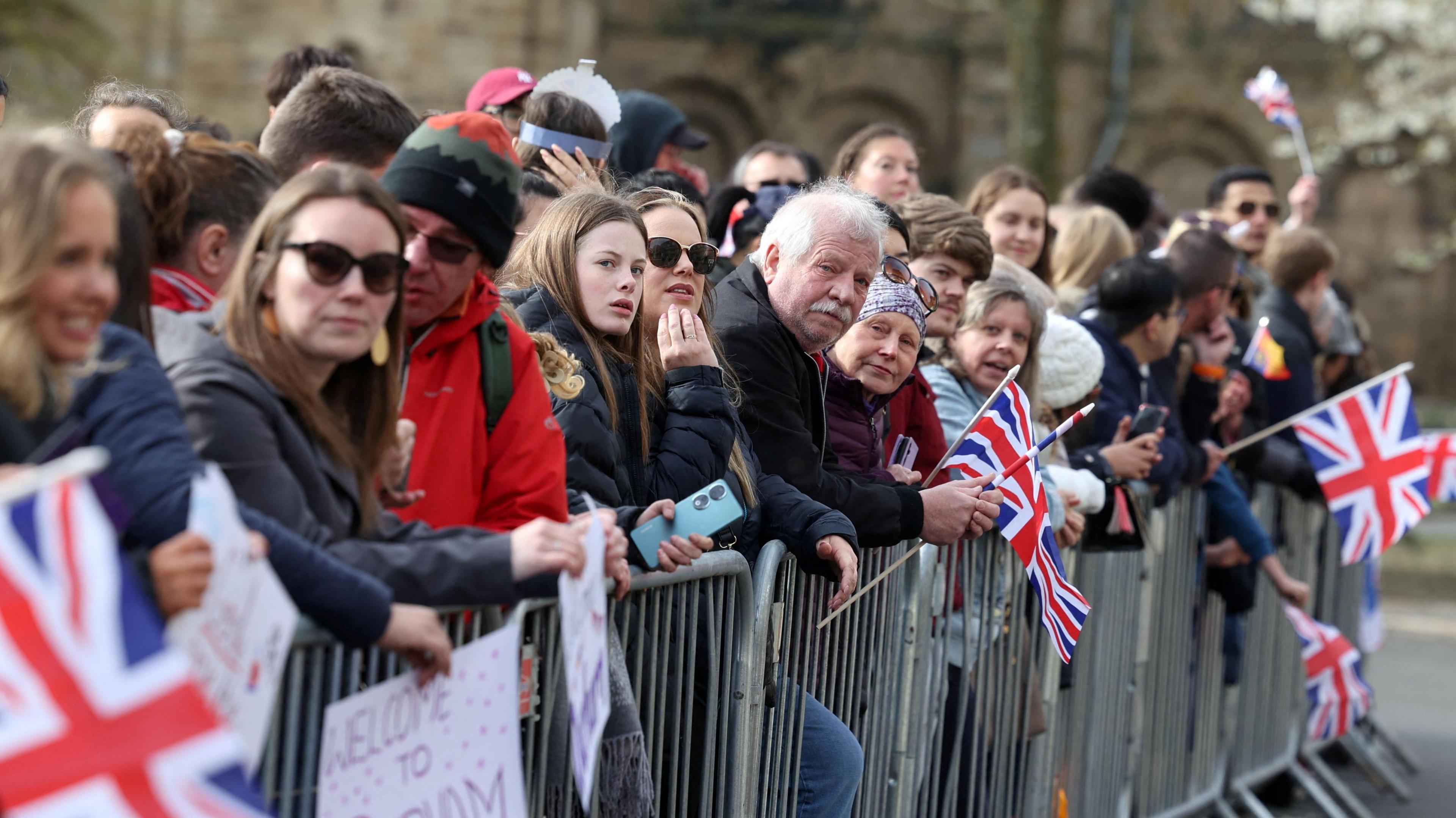 People awaiting the Royal arrival, pressed up against a fence waving Union Jacks. One woman holds a sign reading 'Welcome to Durham'.
