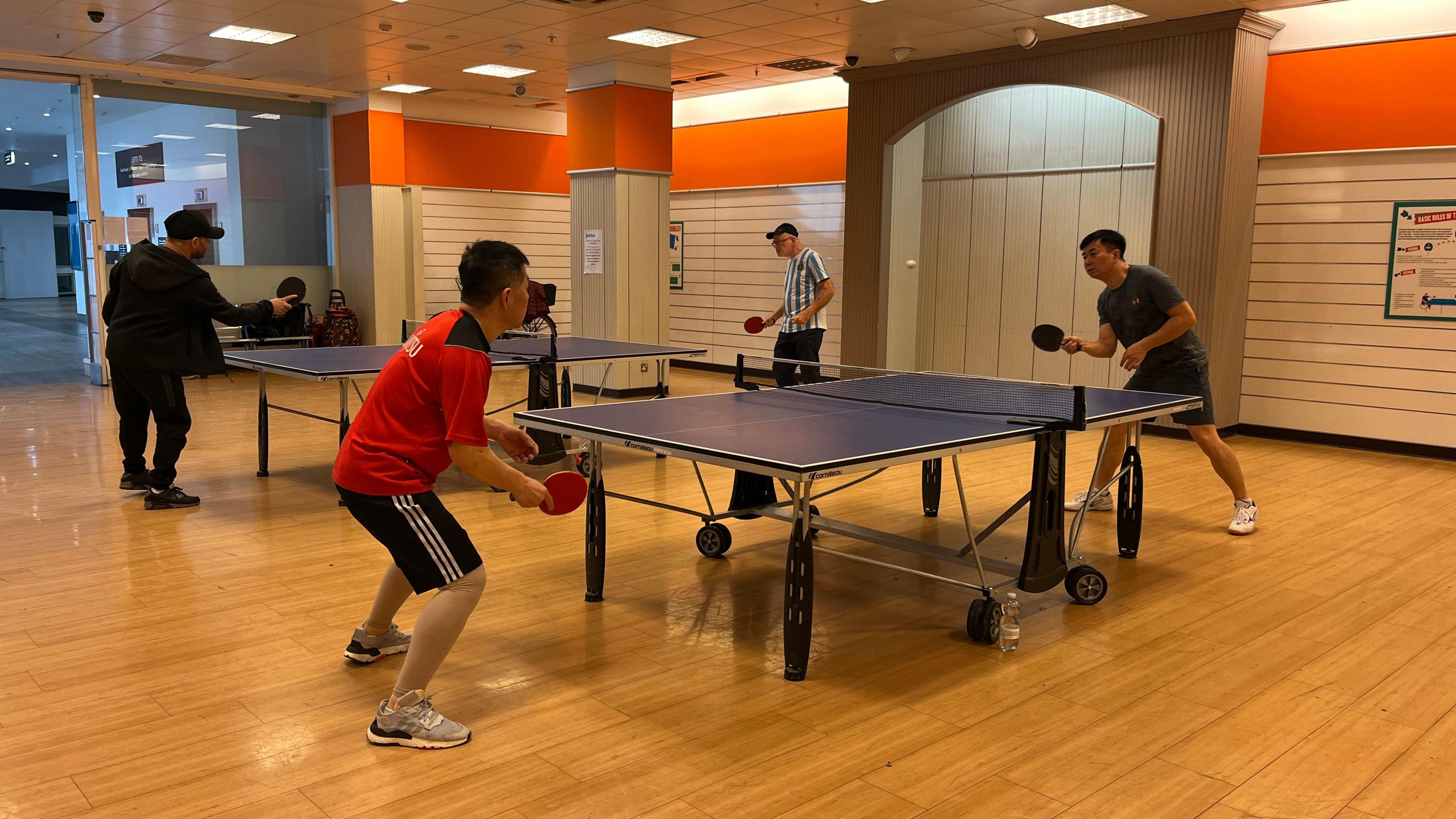 Four men are playing table tennis in an open space with a wooden floor.