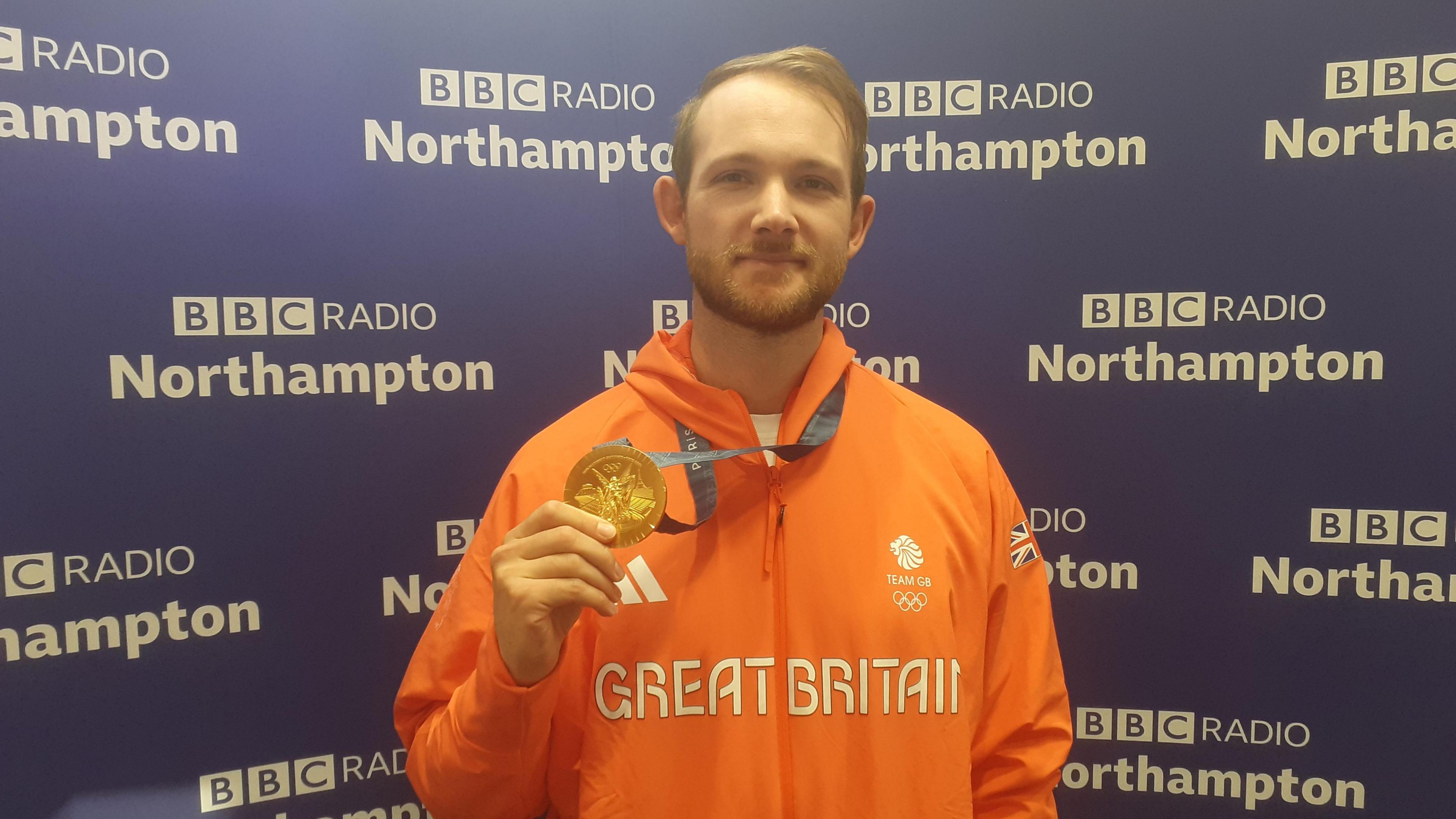 A man in an orange Team GB tracksuit holds up his gold medal