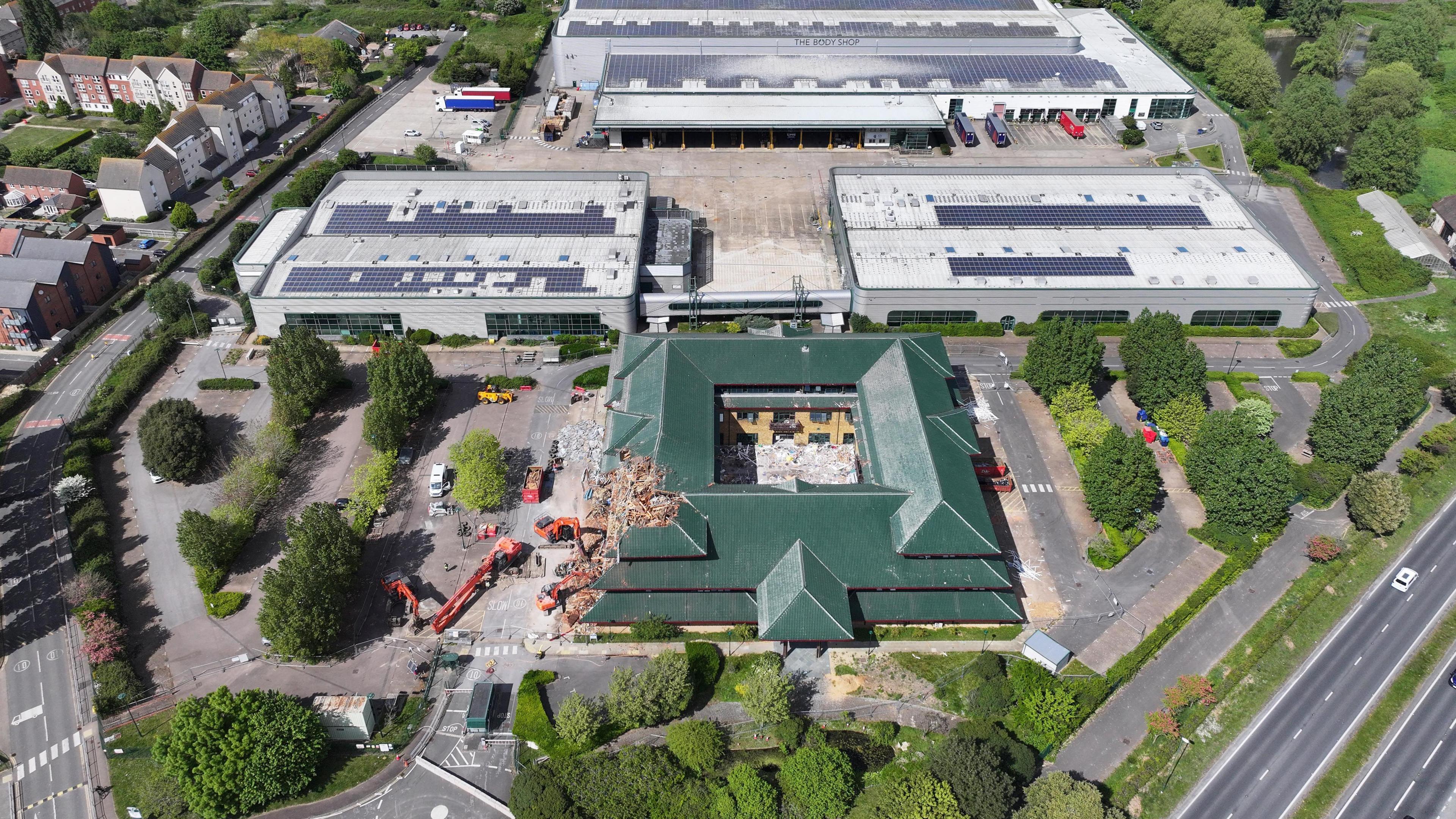 An aerial image of demolition works on a square building with a dark green roof. The building is surrounded by cranes and has a chunk taken out of its side. the roof has a number of triangular layers.