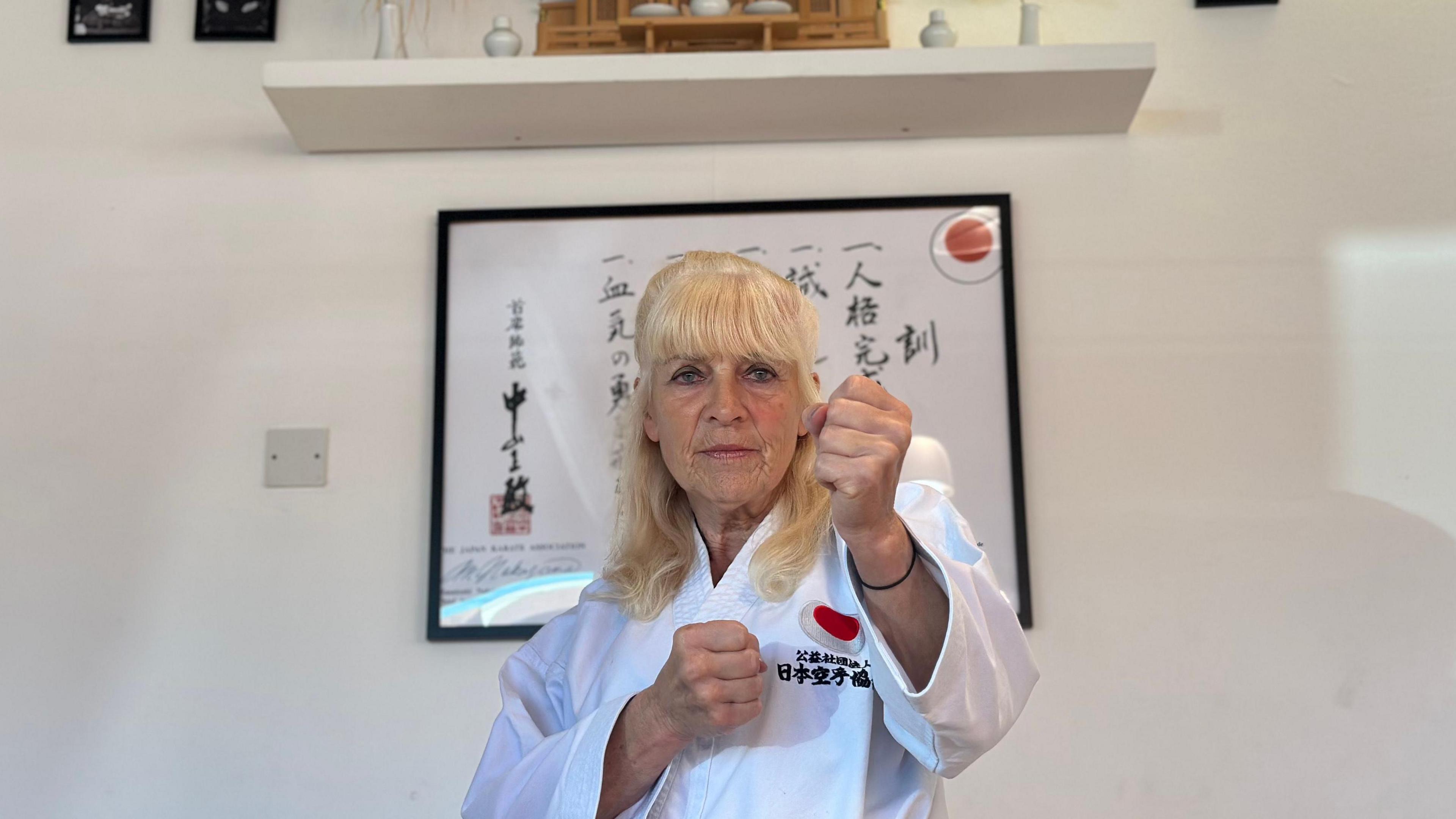 Janet Farrell does a "punching" pose - on this occasion she is facing the camera. She has blonde hair and is wearing traditional white robes