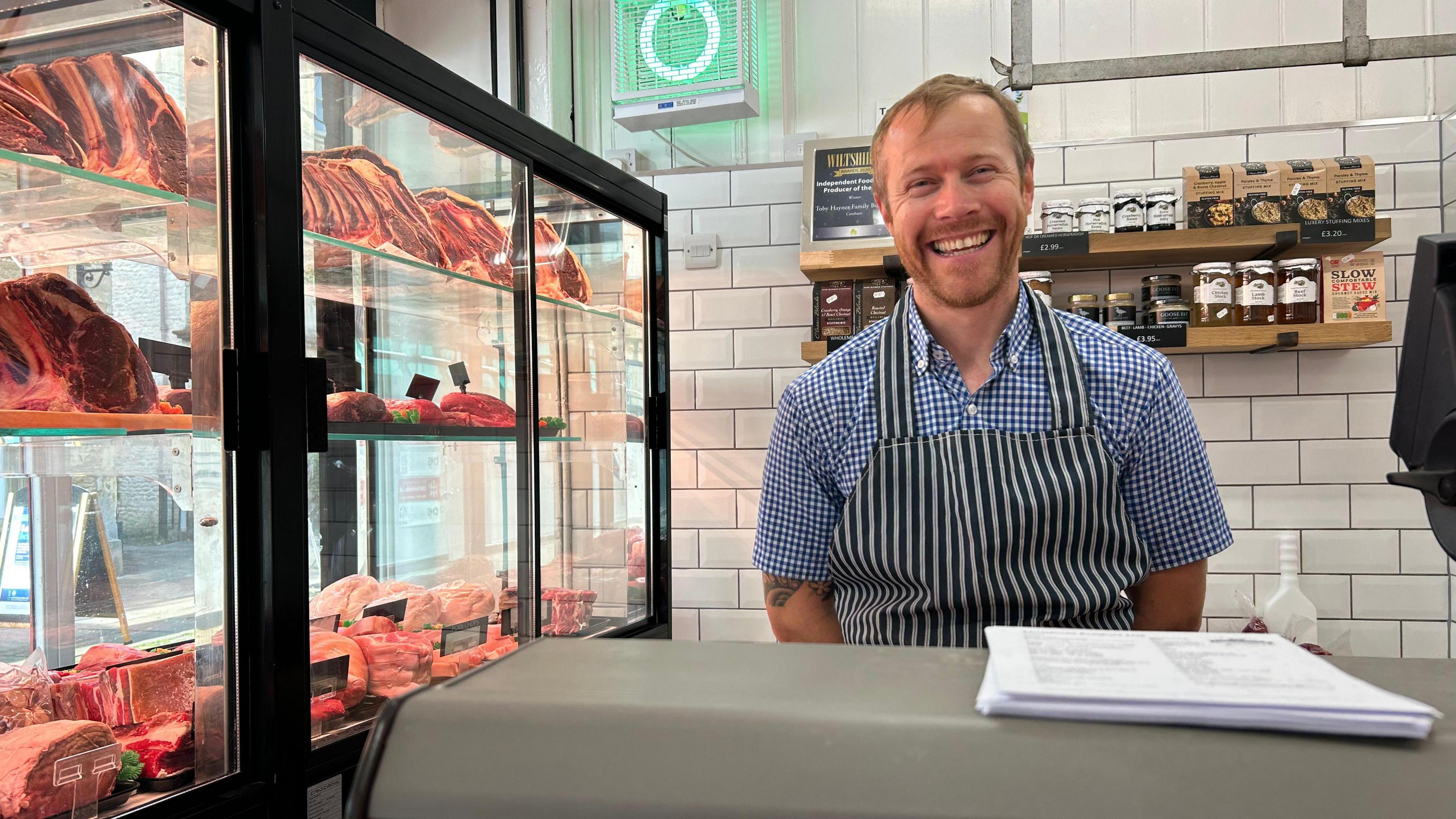 Toby wearing butcher's overalls inside his shop with meat in a glass window cabinet to his left. He is smiling at the camera