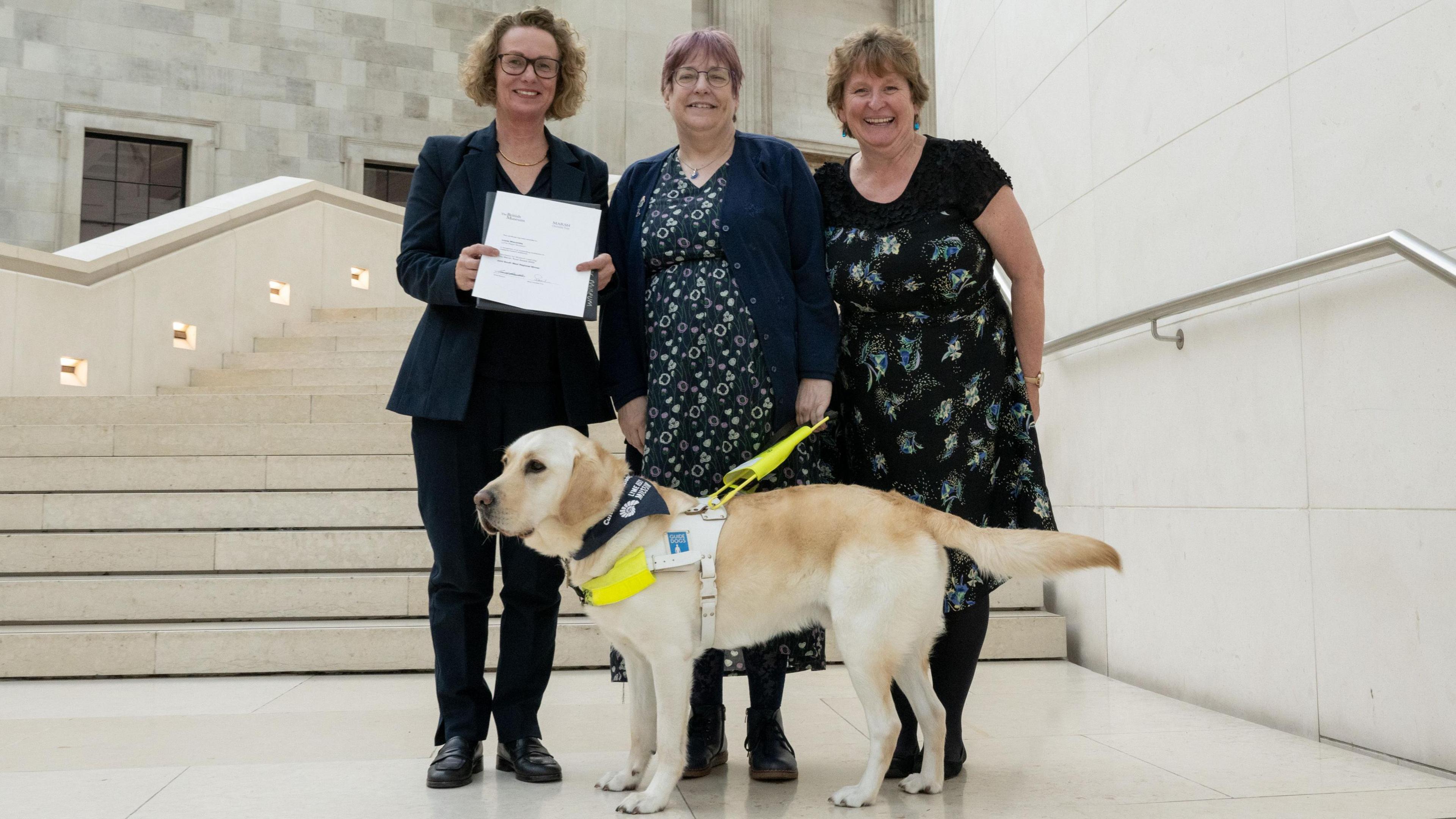 Bridget Houseago, Lizzie Wiscombe, Lin Paterson and Baden the guide dog posing for the camera on the white stone steps of the British Museum's Great Court