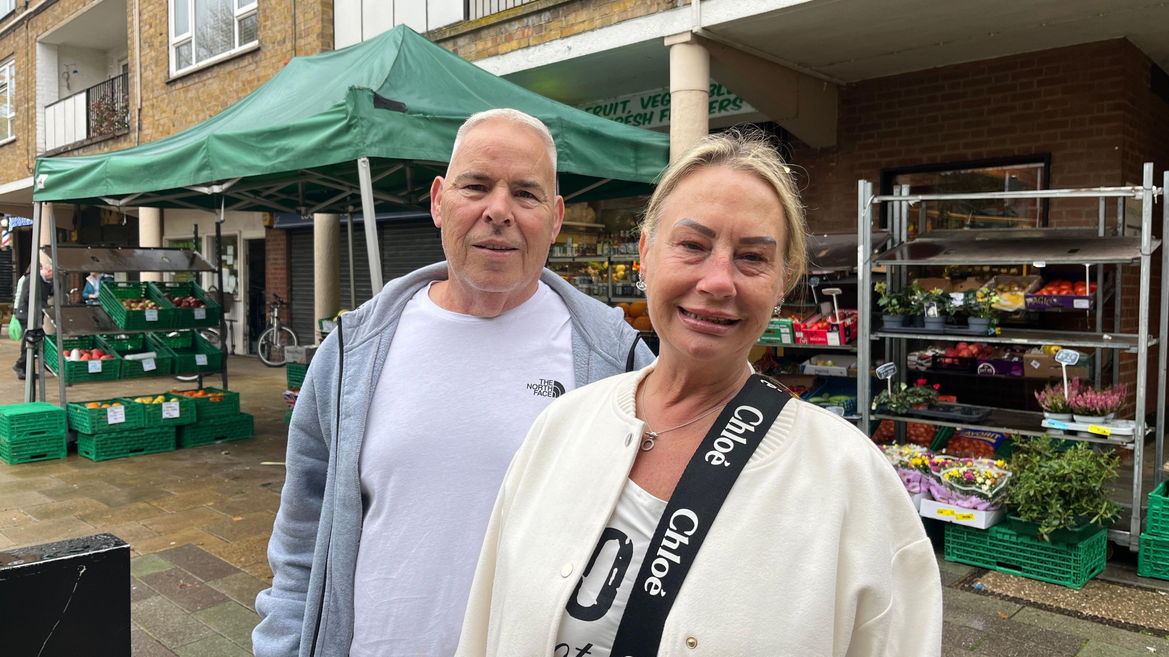 Malcom Everett and Karen Newman are standing in front of a market stall shaded by a green canopy. The stall is filled with fresh produce and flowers arranged in green crates and metal racks. Behind them is a brick building with balconies and windows. Mr Everett is wearing a light jacket with a 'Chloé' branded bag strap, while the other is dressed in a grey hoodie layered over a white t-shirt.