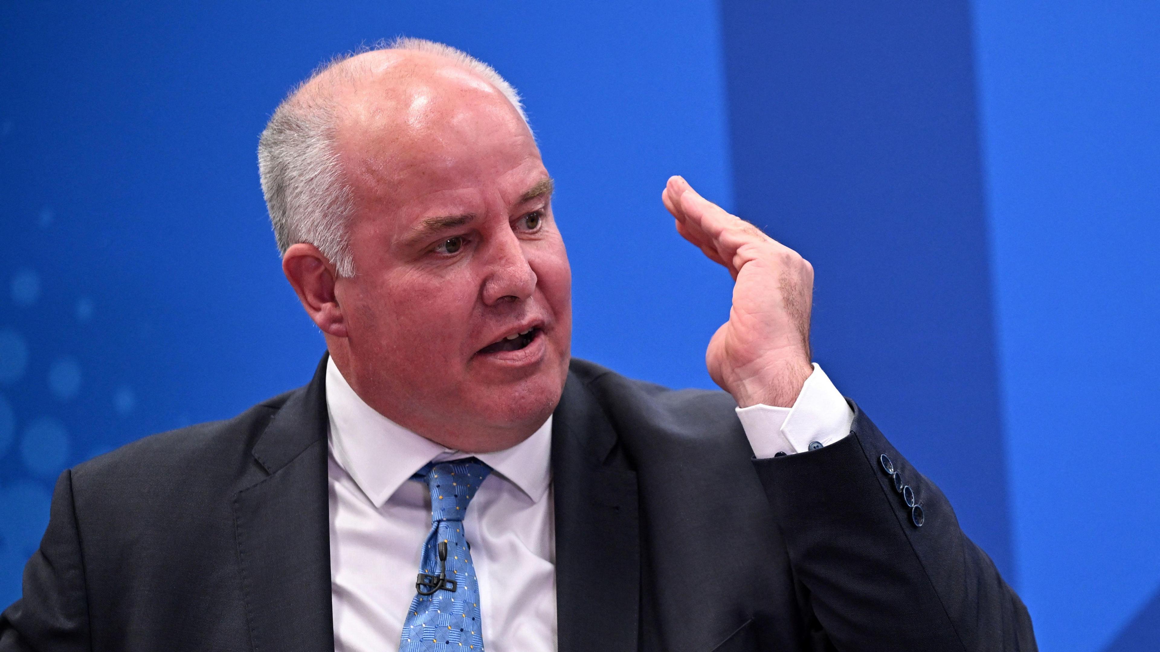 Andrew RT Davies in a suit with a blue tie speaking in front of a blue background.