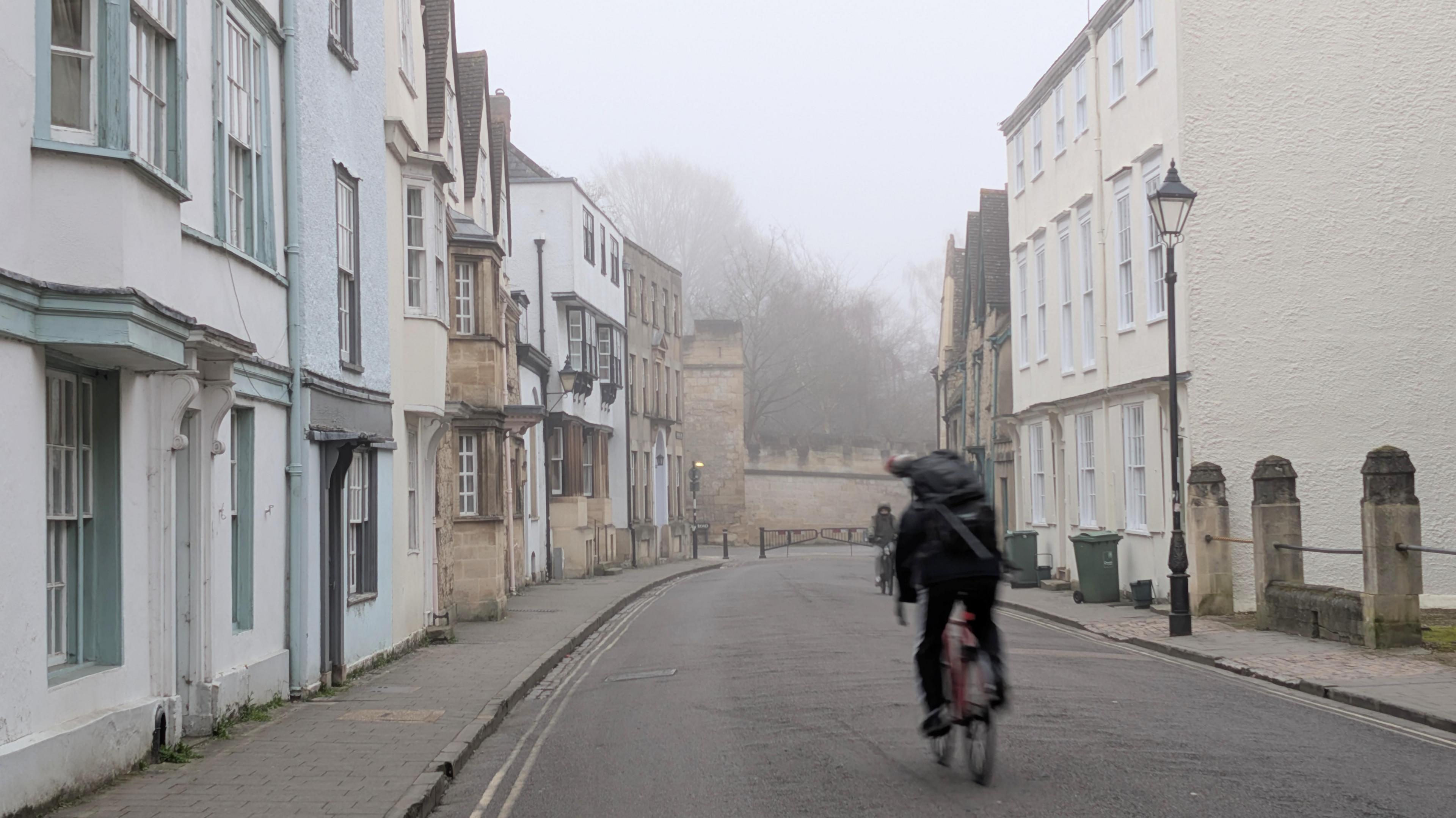 A cyclist in the photo is blurred as they move at speed through the shot. The street is lined with houses of various colours. The scene is foggy and you can't see the sky. Trees appear through the murk behind a low wall.