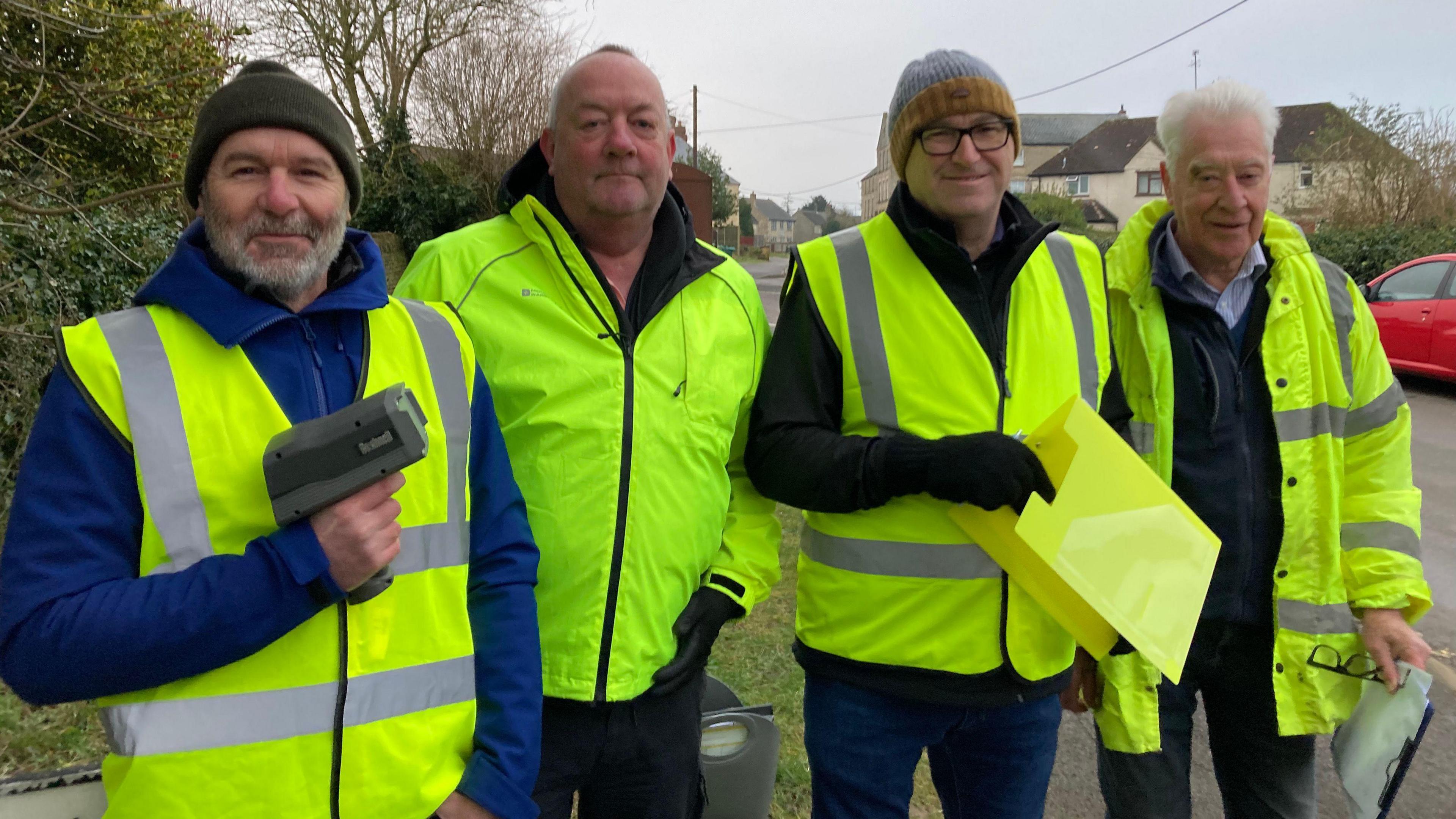 Four men in aged somewhere between 50-80, all wearing high-visibility clothing, pose shoulder to shoulder by the side of a residential road. The man on the far left is holding up a small camera, which resembles a checkout gun. Another man is holding a clipboard