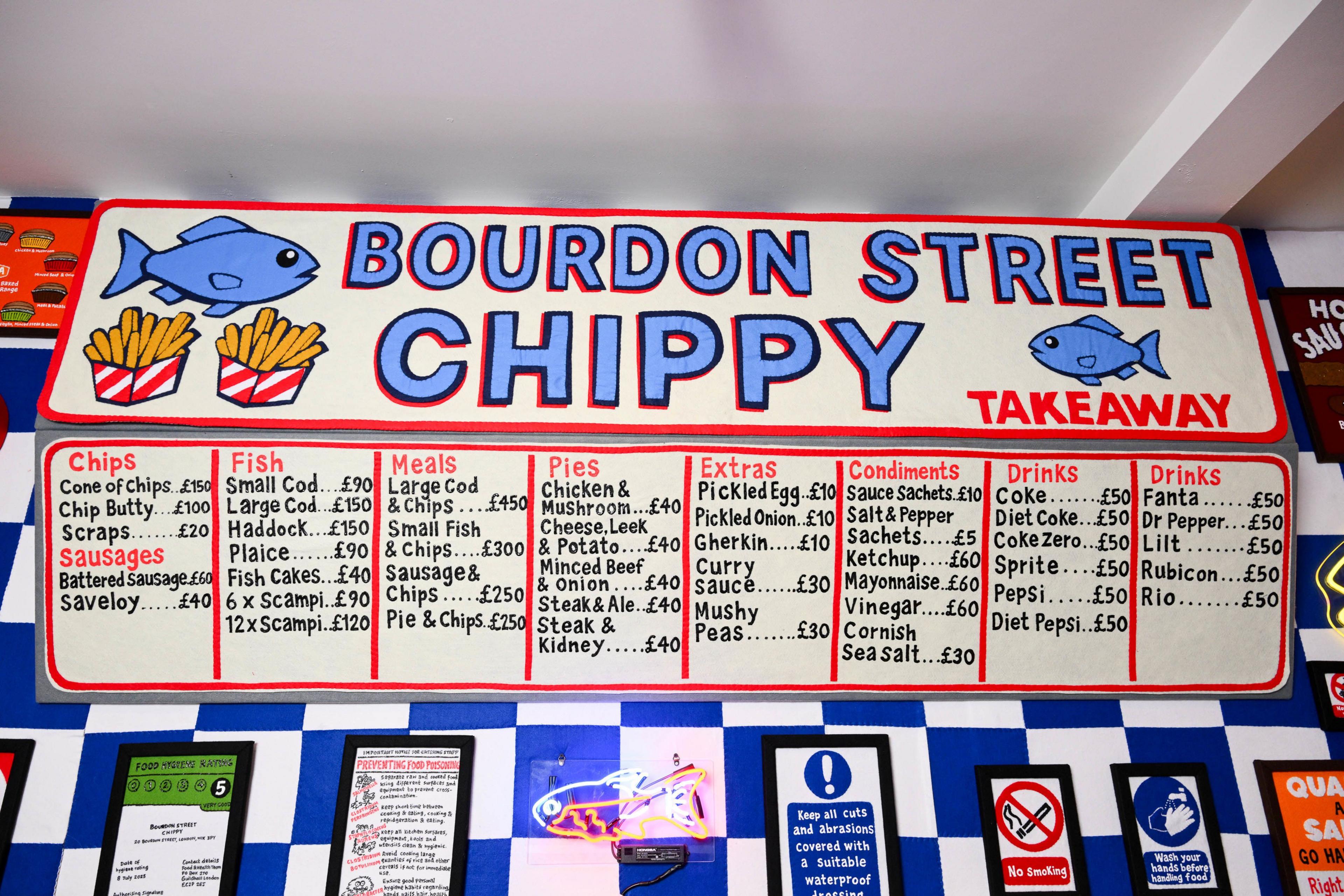 Image of large chip shop menu that reads Bourdon Street Chippy, behind the counter. The wall is blue and white, and has lots of different signs on it.