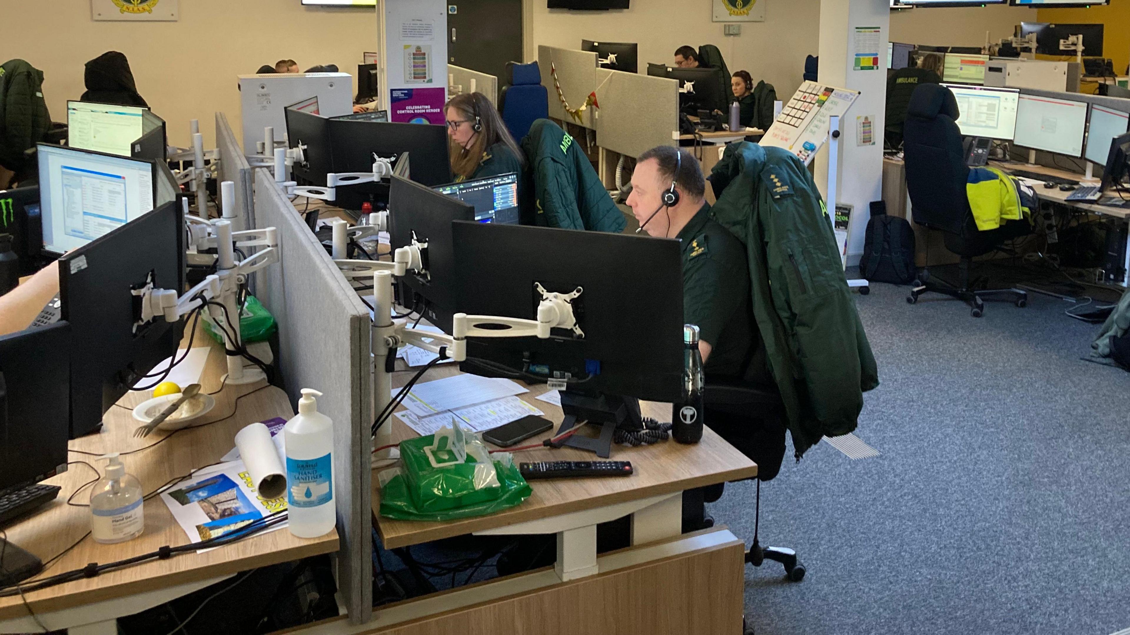 NIAS control room. People in ambulance uniforms are sitting at desks in front of computer screens wearing headsets.