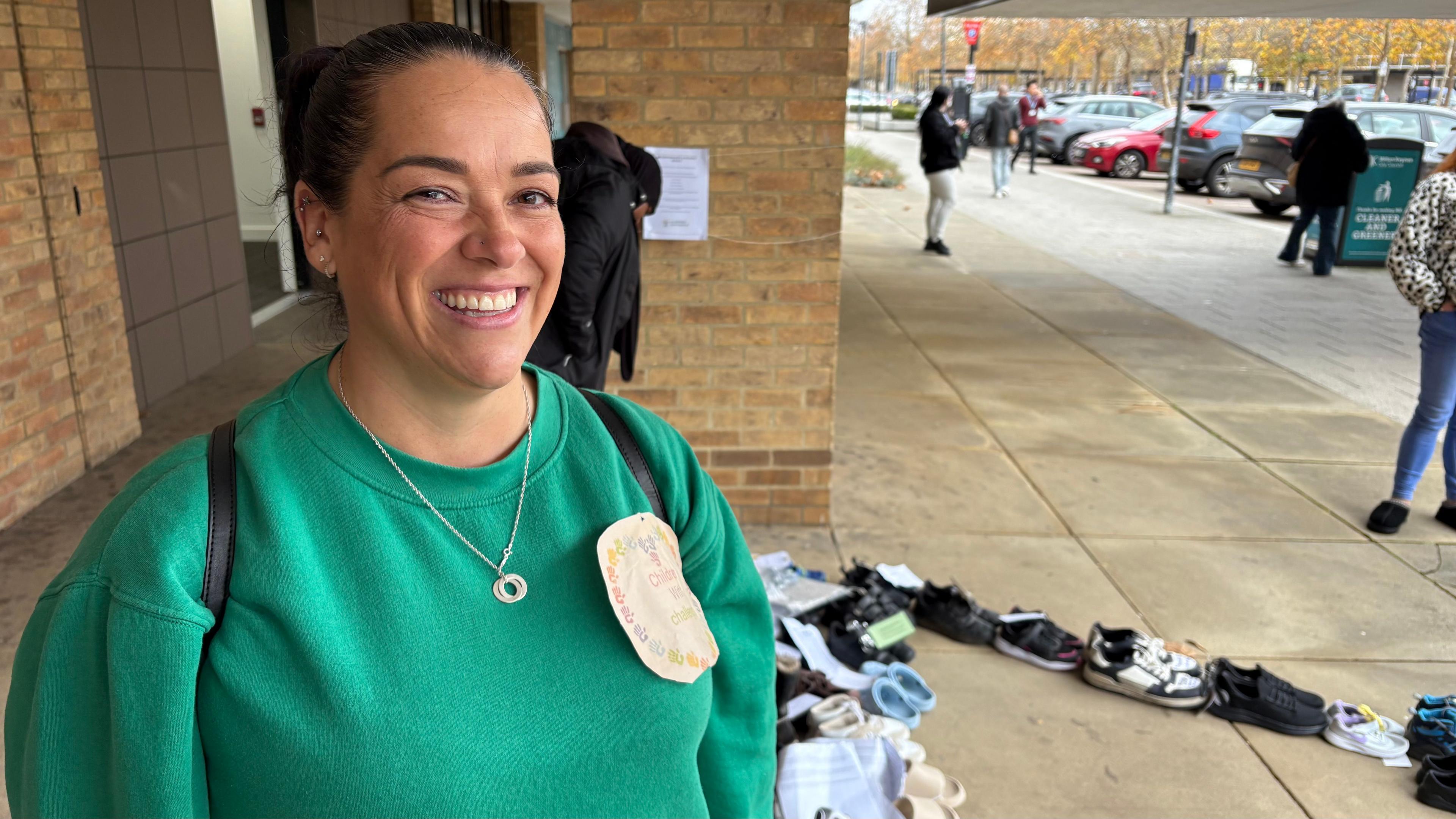 Amanda, who has short dark hair, smiling at the camera and wearing a small silver necklace. She has a large square badge attached to the green top she is wearing. There is a line of shoes to her left and she is standing in front of a brick-built building. There is a pavement to the right of the picture.