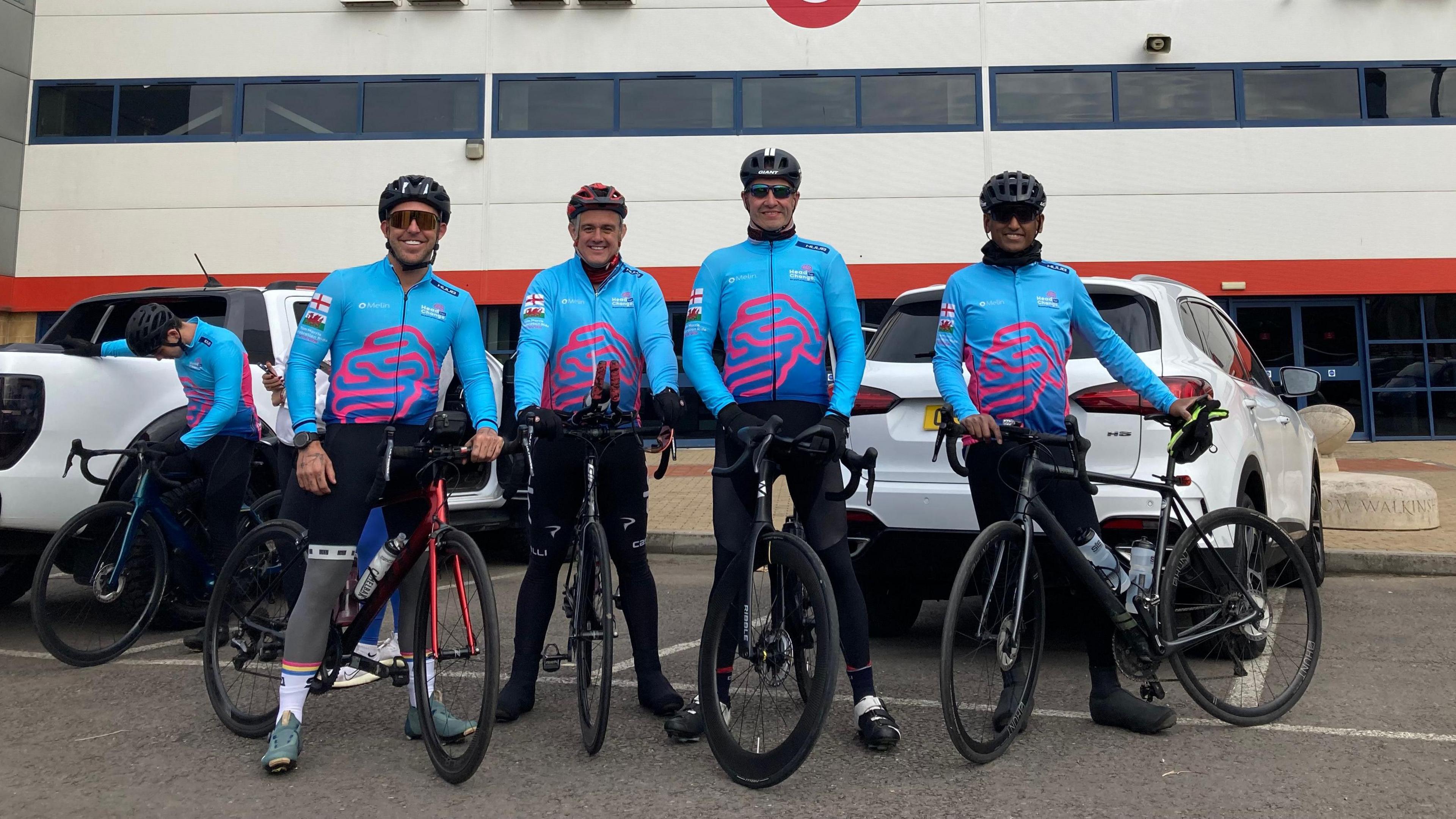 Four men wearing black leggings and matching blue athletic shirts, sunglasses and bike helmets. They are standing in front of Gloucester Rugby Club, posing with their bikes. 