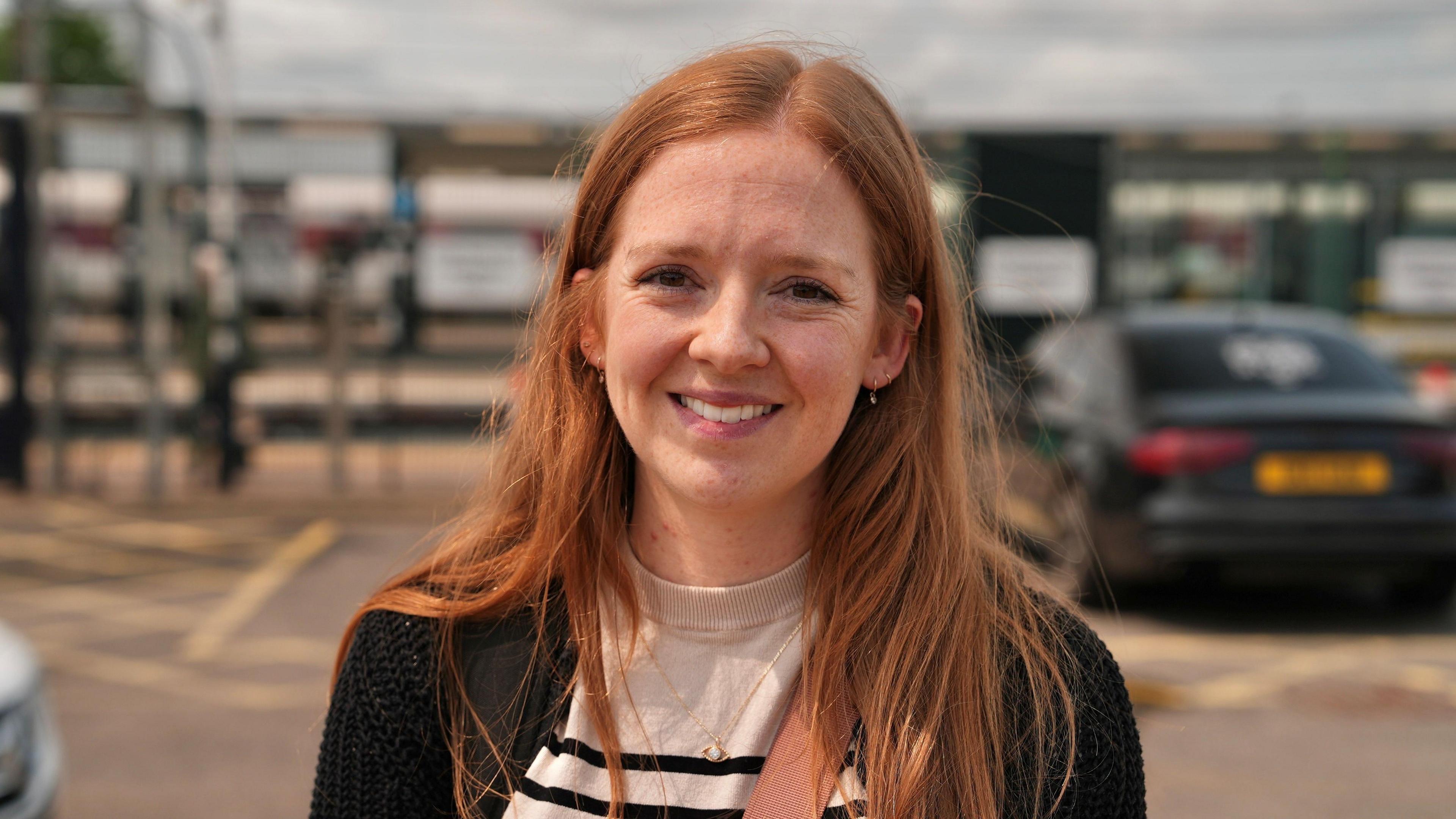 Victoria outside Peterborough station, in the background are cars parked in the car park. She has red hair and is wearing a black jacket with a black-and-white striped T-shirt underneath.