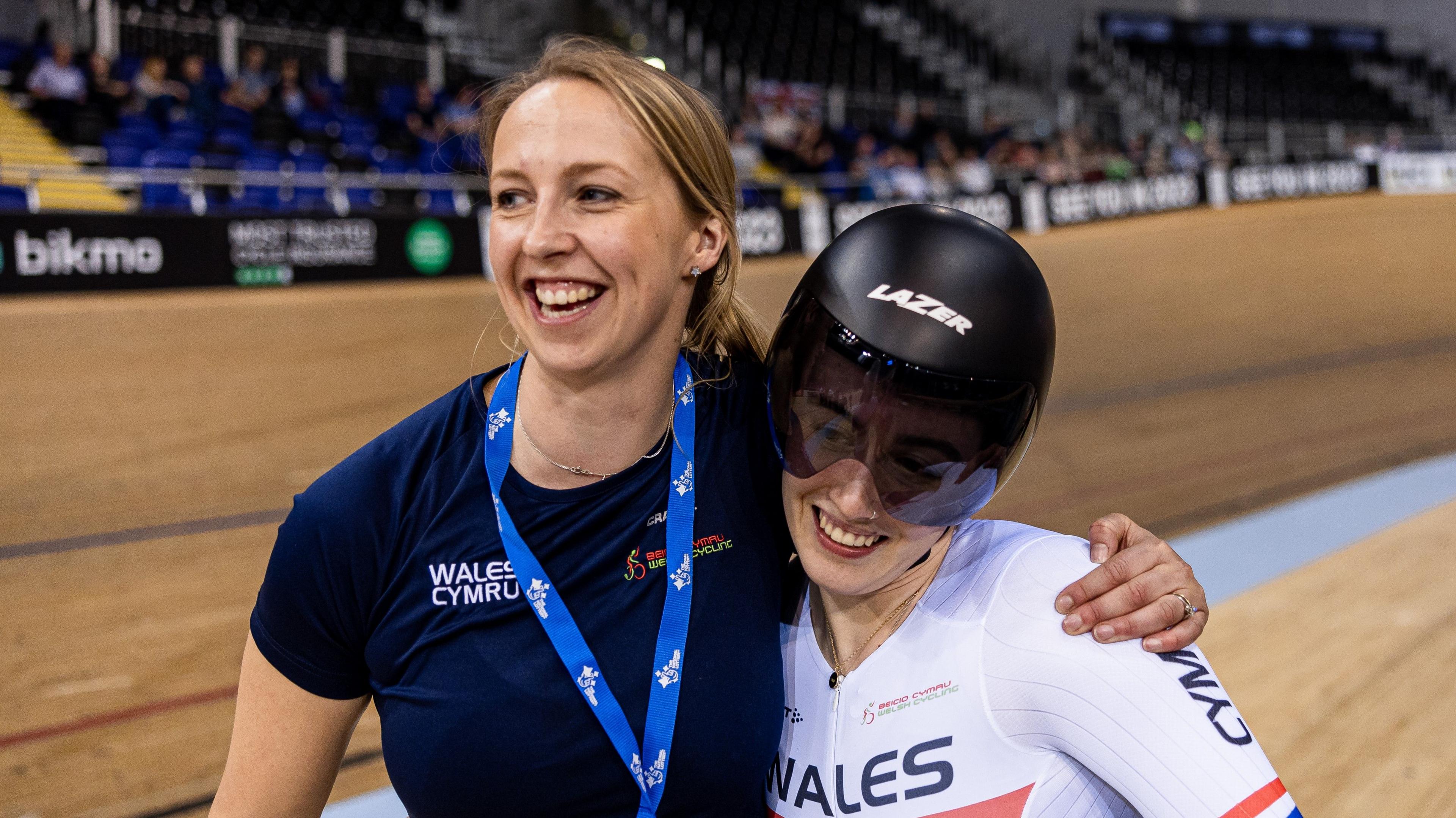 Rachel Draper with her arm around a smiling Lowri Thomas who's in Welsh cycling kit and wearing a track helmet. They're next to a cycling track. Rachel has her blonde hair tied back and is wearing a blue lanyard and navy t-shirt.