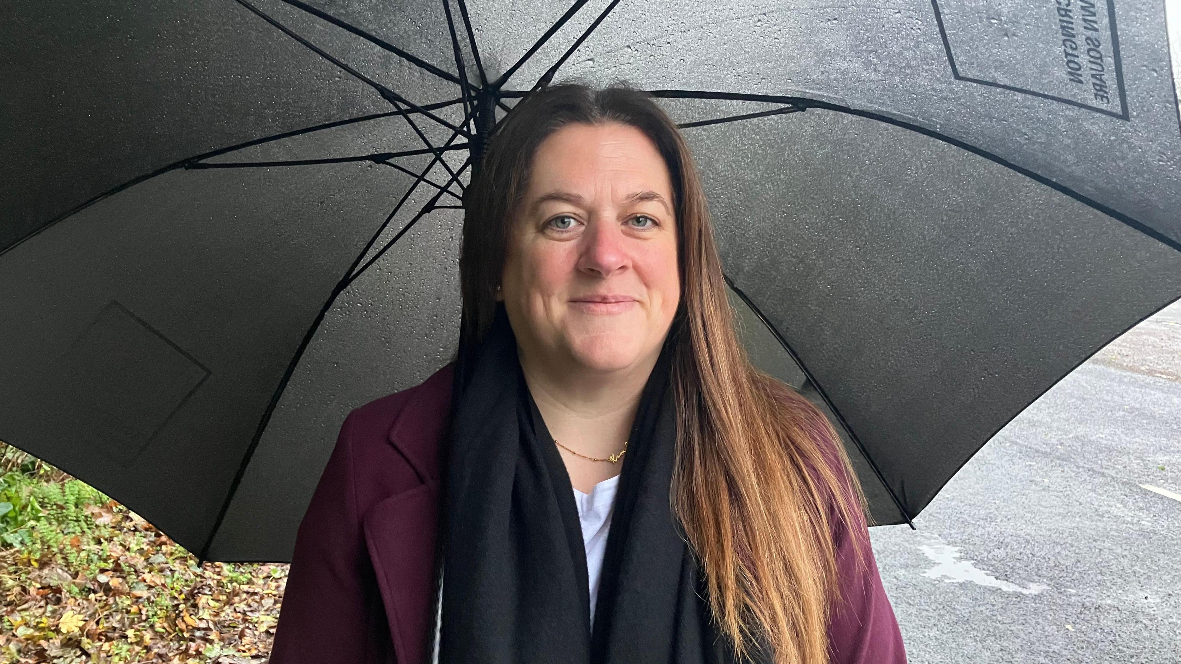 Kimberley Whitehead is pictured wearing a purple blazer and navy scarf. She is standing next to a road on a rainy day and is underneath a large black umbrella.