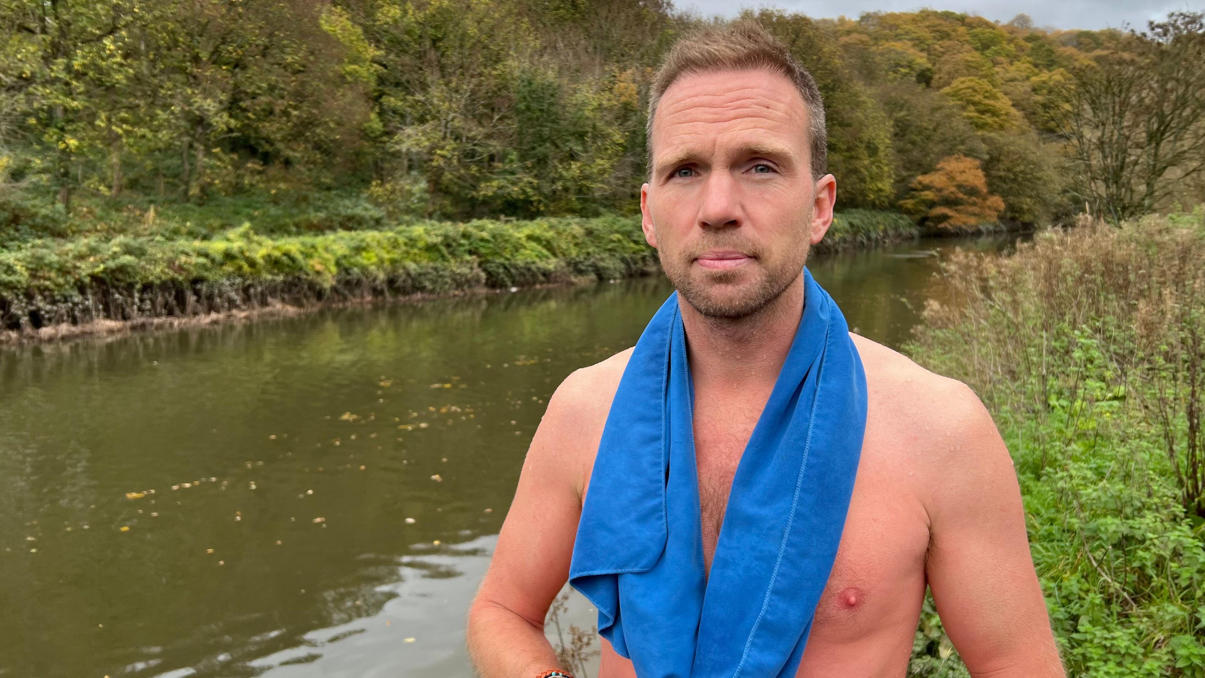 Johnny Palmer stands on the bank of the River Avon after a swim. His top half is visible and he is only has a towel around his shoulders. 