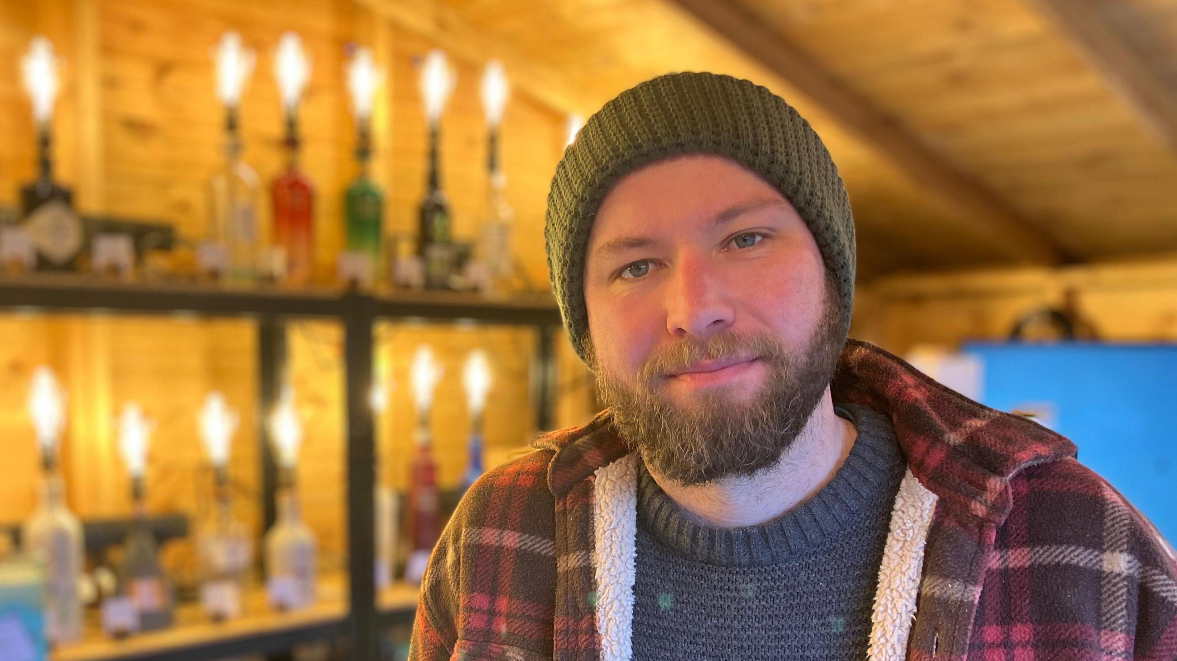 Nicholas Hocking wearing a green beanie and a check jacket looks at the camera. He has a brown beard and is stood inside a log cabin.