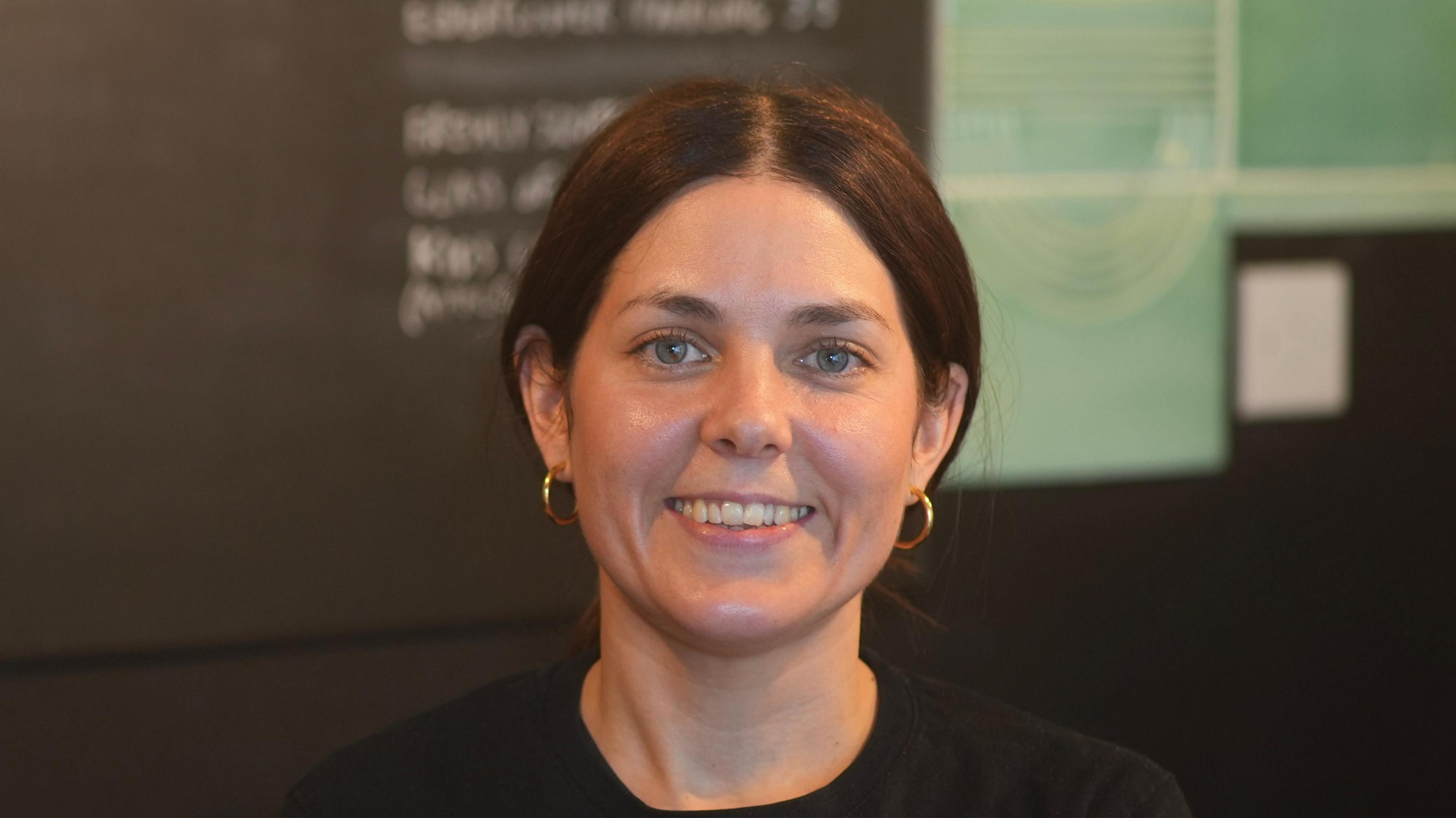 A woman with brown hair that is tied up behind her head smiles at the camera while standing in a coffee shop. She has gold hoop earrings and is wearing a black long sleeved top.