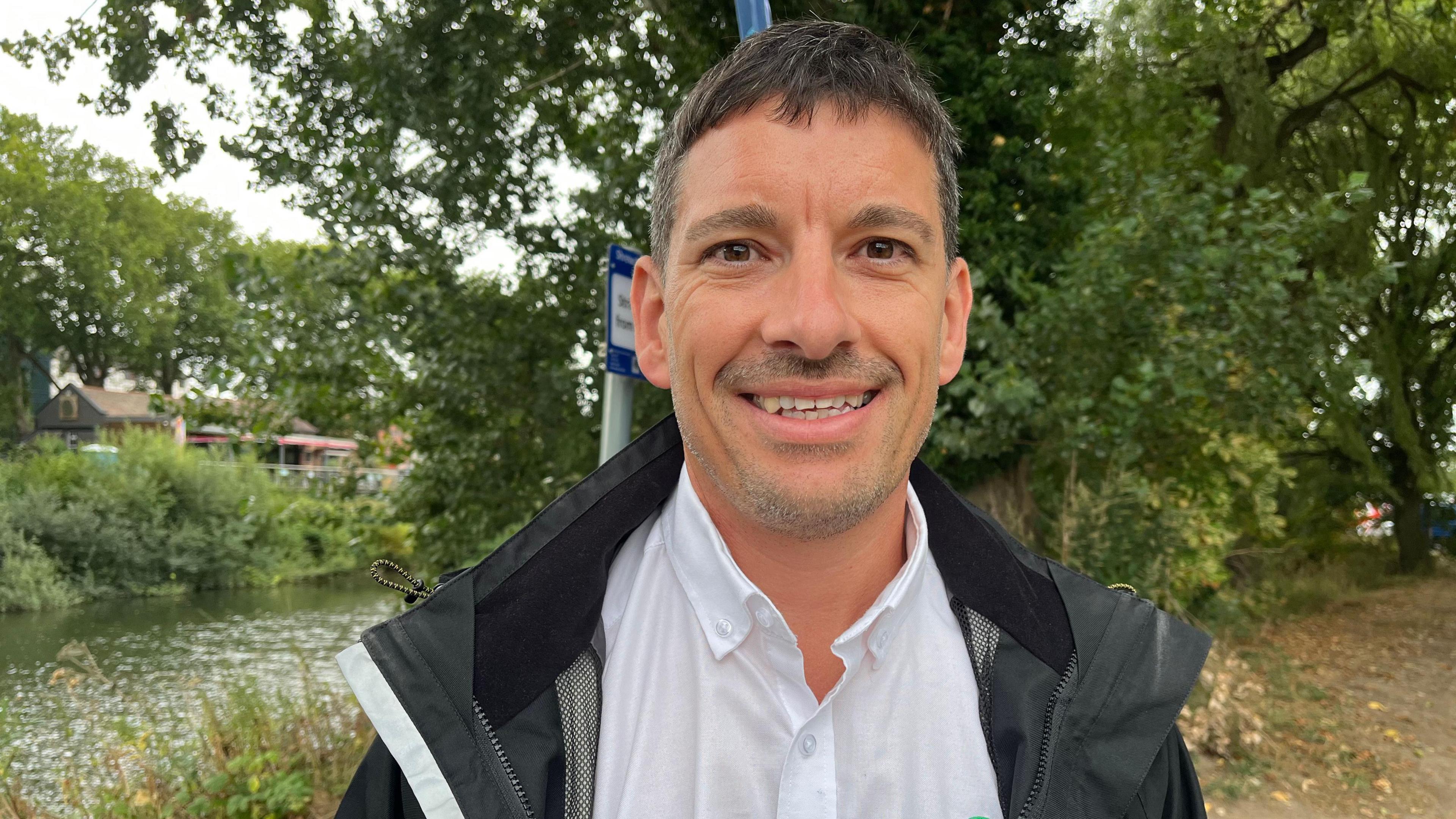 Martin photographed looking into the camera and smiling - he has short dark hair and is wearing a white collared shirt and a black waterproof coat. Behind him you can see the Severn, which is lined by trees and grasses.
