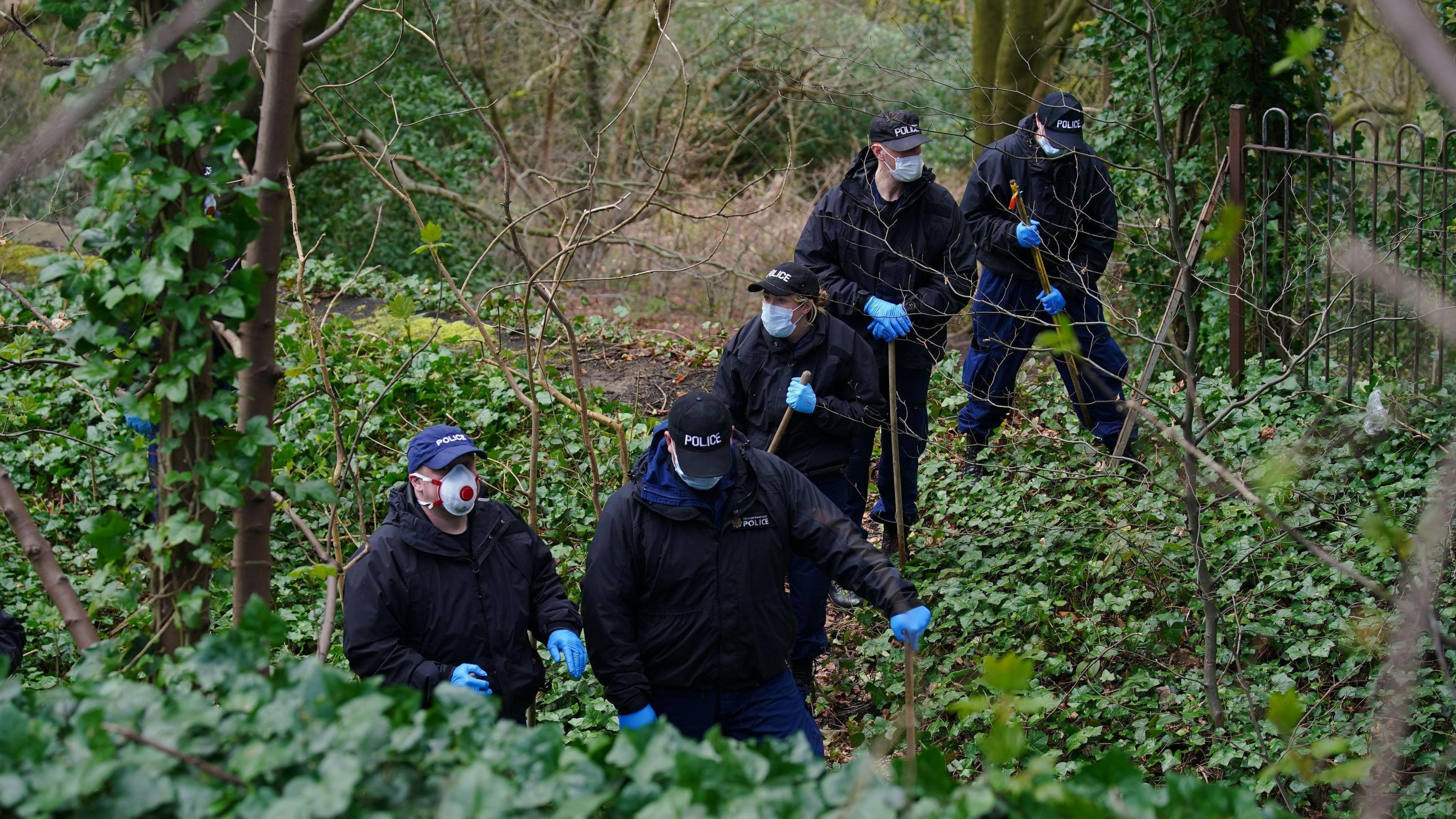 Police officers wearing medical face masks and carrying sticks in shrubbery - they are wearing blue gloves and in a line - their caps read Police