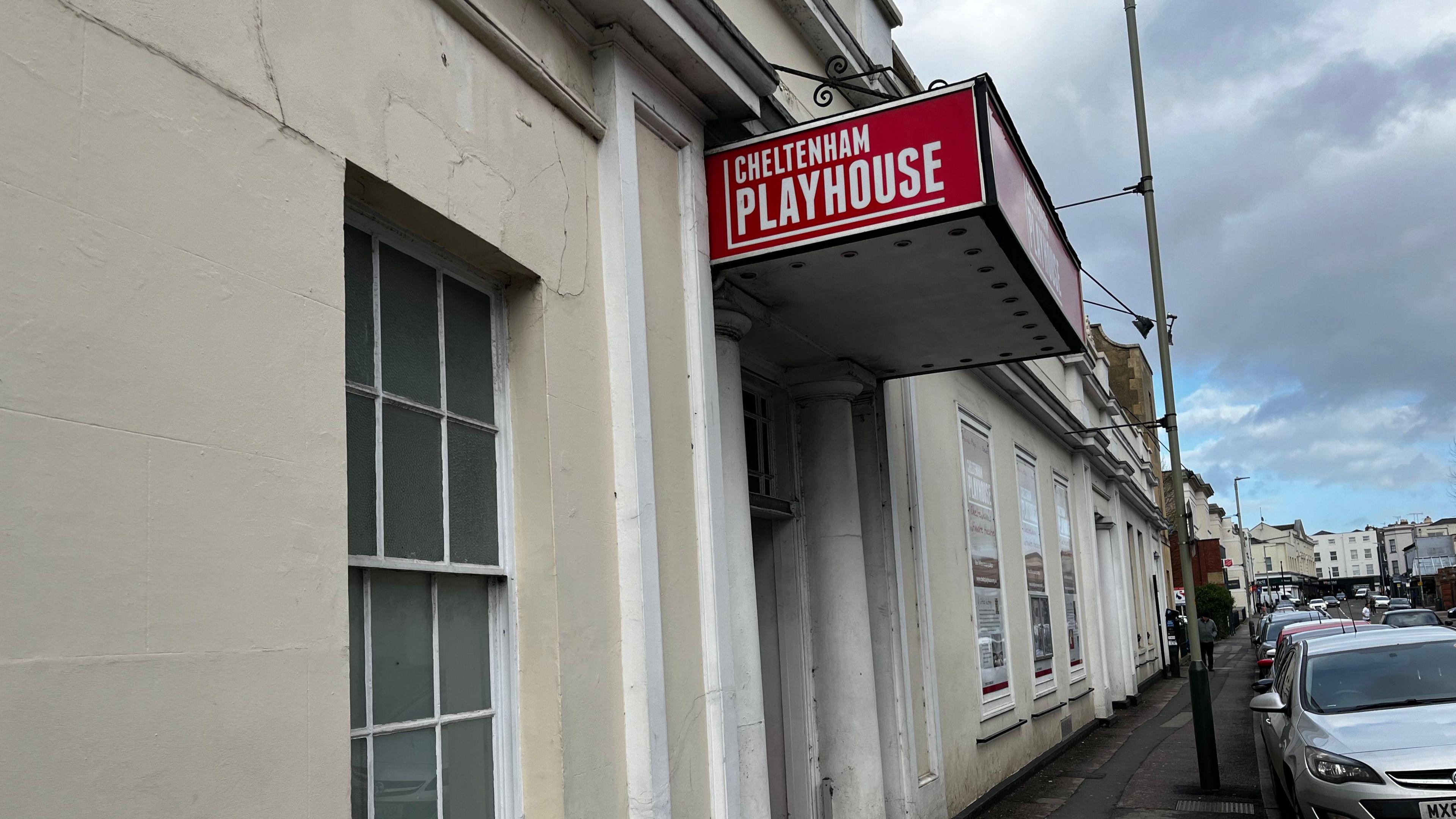 A white Regency style building with a large red sign coming out of it over its door which reads "Cheltenham Playhouse". Cars are parked on the road outside and more buildings can be seen in the distance.