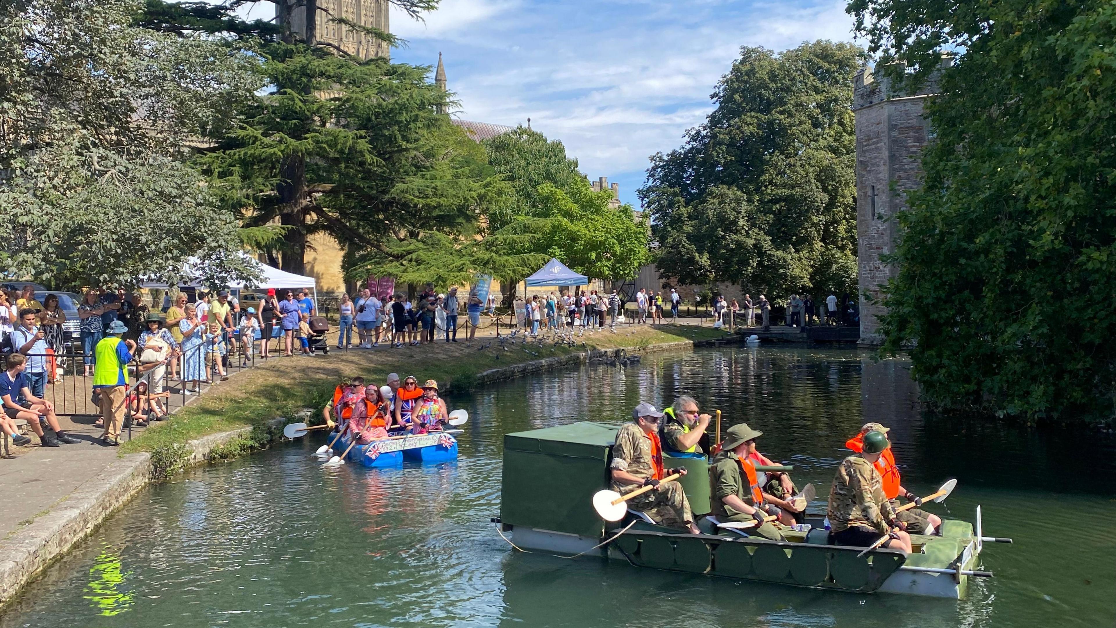 Two different teams are in the water of the moat at Wells Bishop's Palace as they prepare for the annual raft race. One team are all in military uniforms and riding a khaki-coloured craft. The other team are riding a raft sitting on blue barrels. In the background spectators are visible on the bank.
