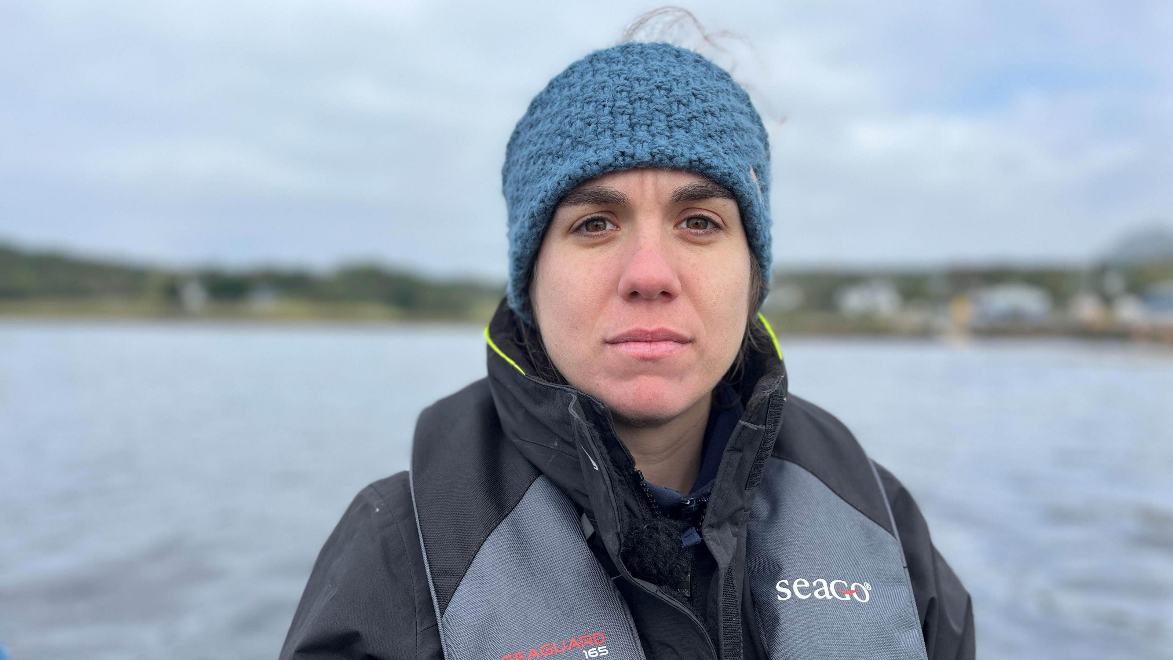 Anna Safryghin wearing a blue woolen head covering, a black jacket and a grey and black lifejacket, with a body of water behind her