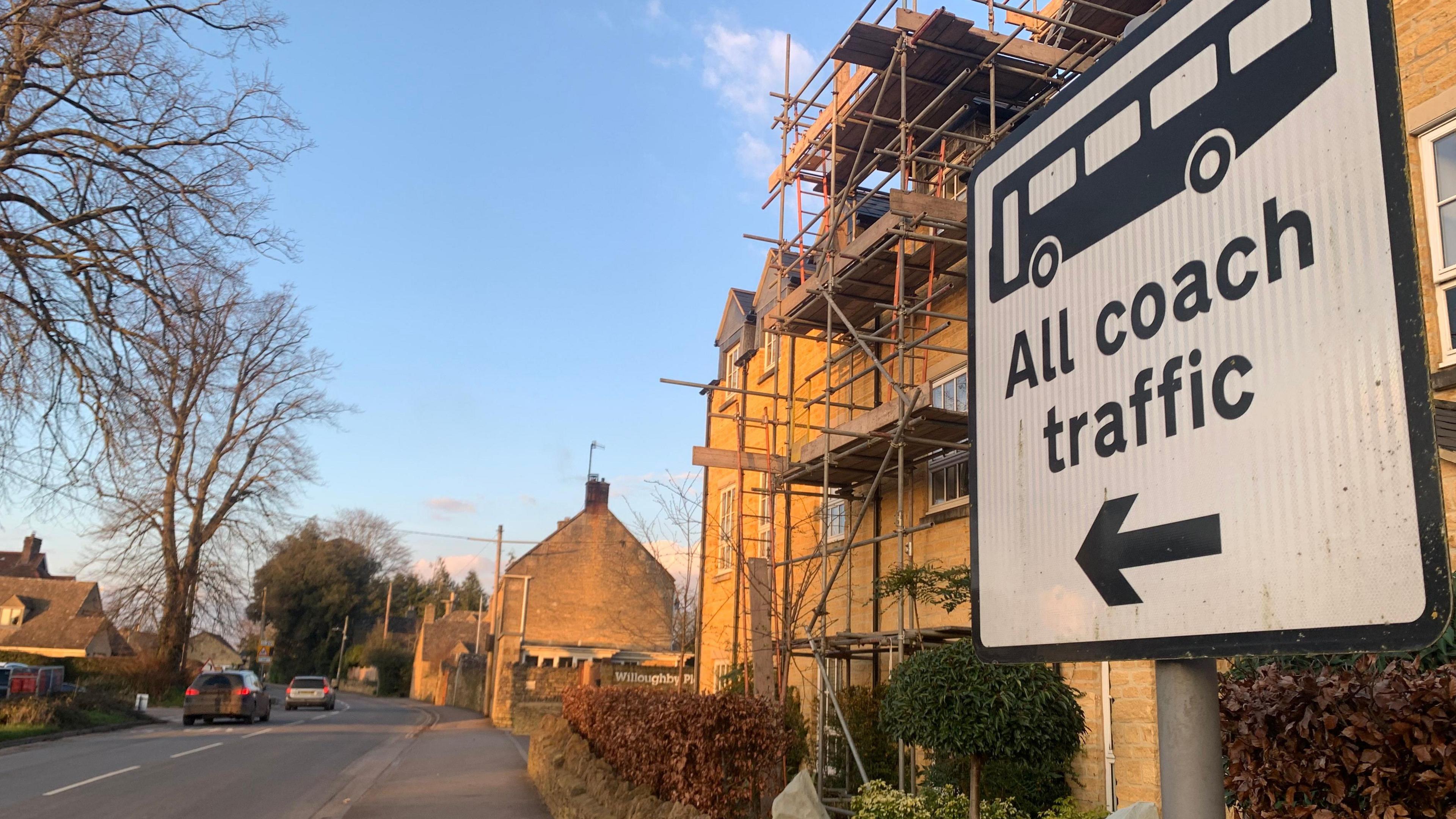 A black and white road sign that reads "All coach traffic" with an image of a bus and an arrow pointing down the road, which is a residential road with Cotswold stone houses, one of which is covered in scaffolding. Two cars can be seen driving down the road.