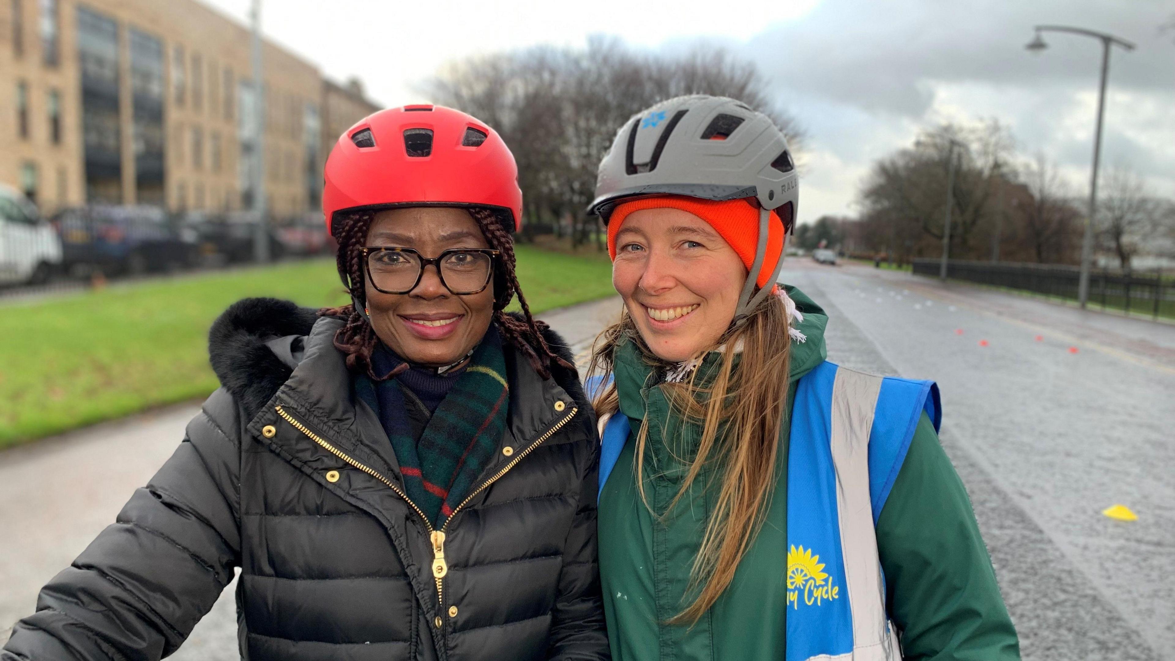 Edith, from Nigeria, and cycling instructor Eilidh. Edith is wearing a red cycle helmet, black puffer coat and tartan scarf. She has long braided hair, tied back. Eilidh has a grey cycle helmet and a green jacket with a bright blue hi-vis vest. She has long, brown hair.